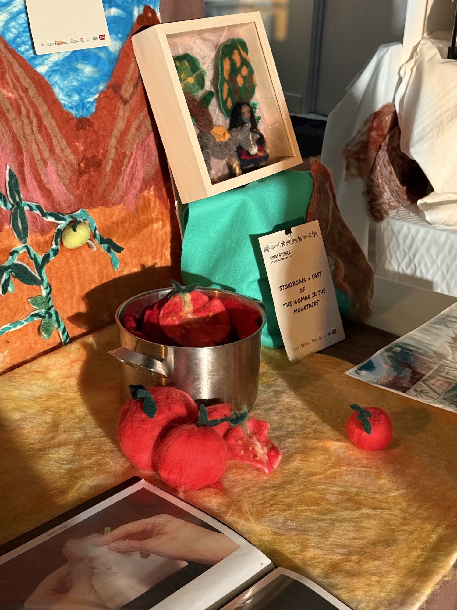 Metal bowl with red contents on wooden table, surrounded by red fabric tomatoes, framed artwork, green objects, and papers.