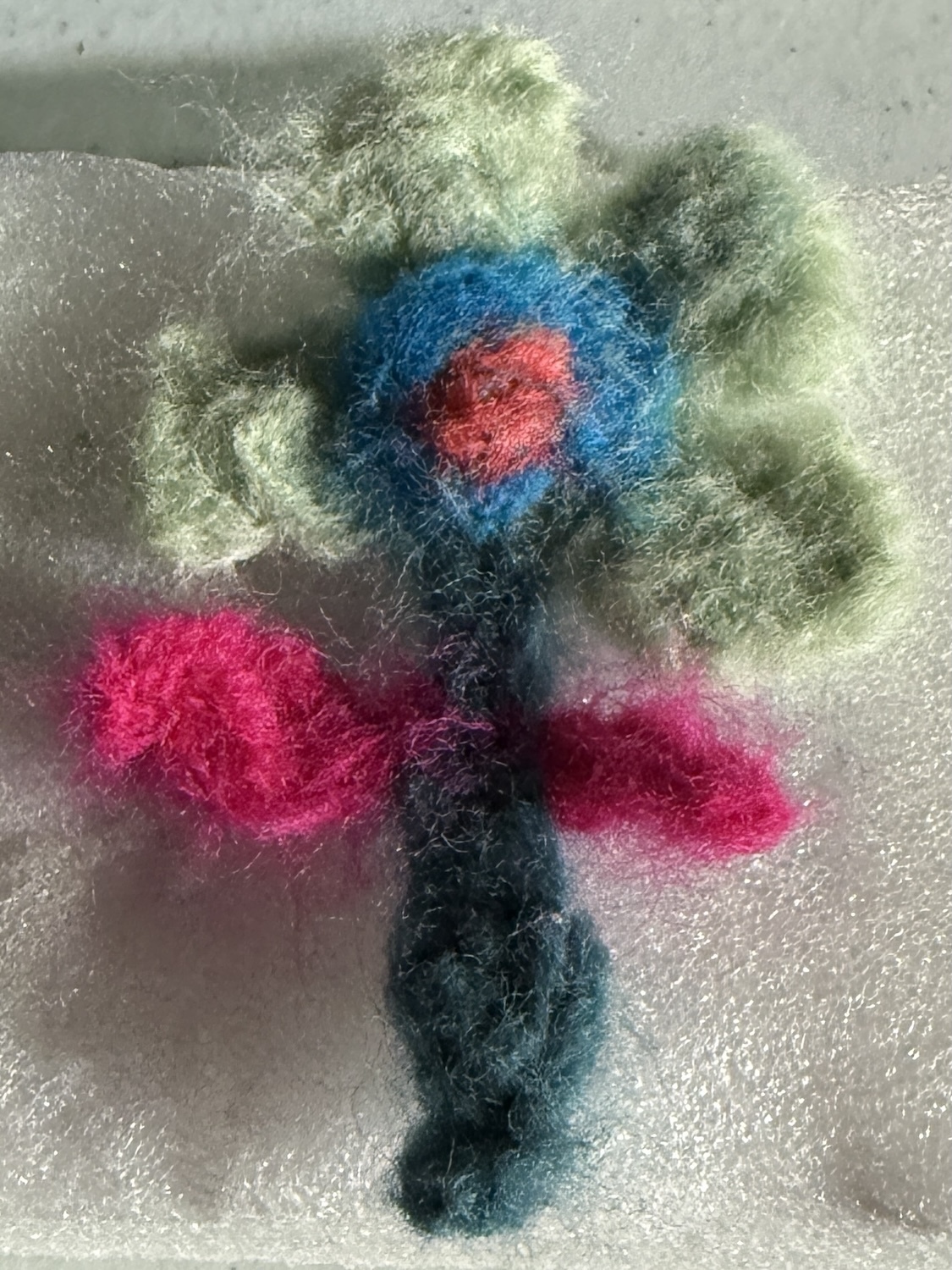 Felted wool flower with green petals, blue centre with red core, magenta side petals, and dark green stem on grey surface.