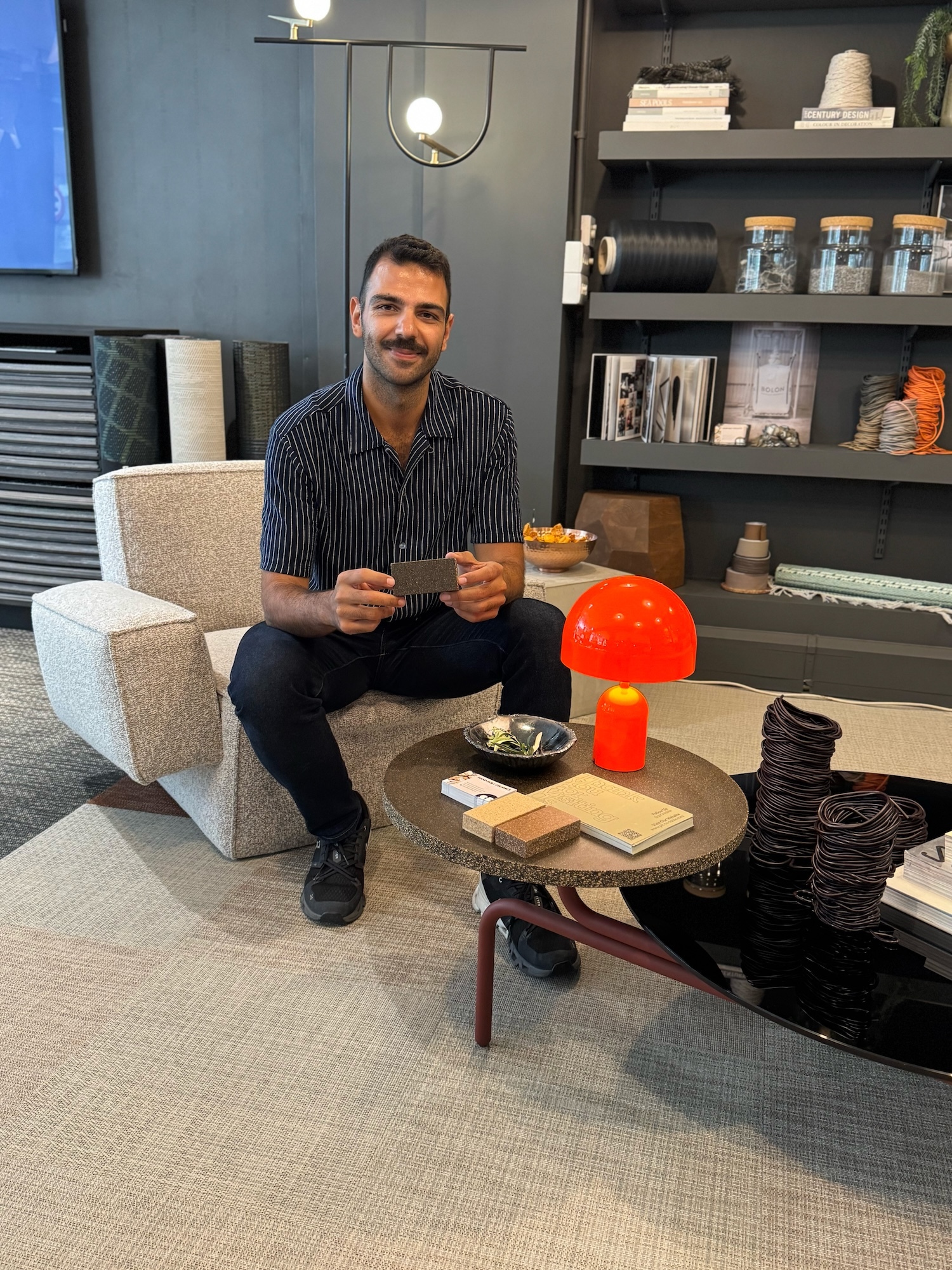 M Afşaroğlu sitting in a boxy white-grey armchair in front of a table made out of sustainable material Pit-Board. He is holding a small sample of the material in his hands.