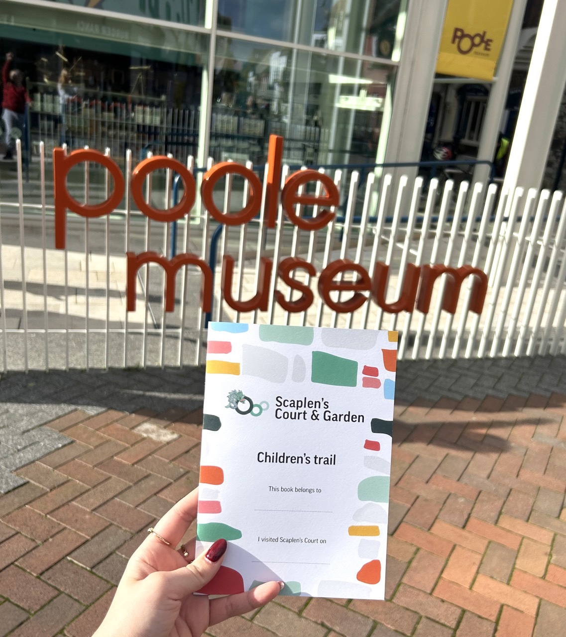 Hand holding children's trail leaflet outside Poole Museum entrance with orange lettering on glass frontage and white railings.