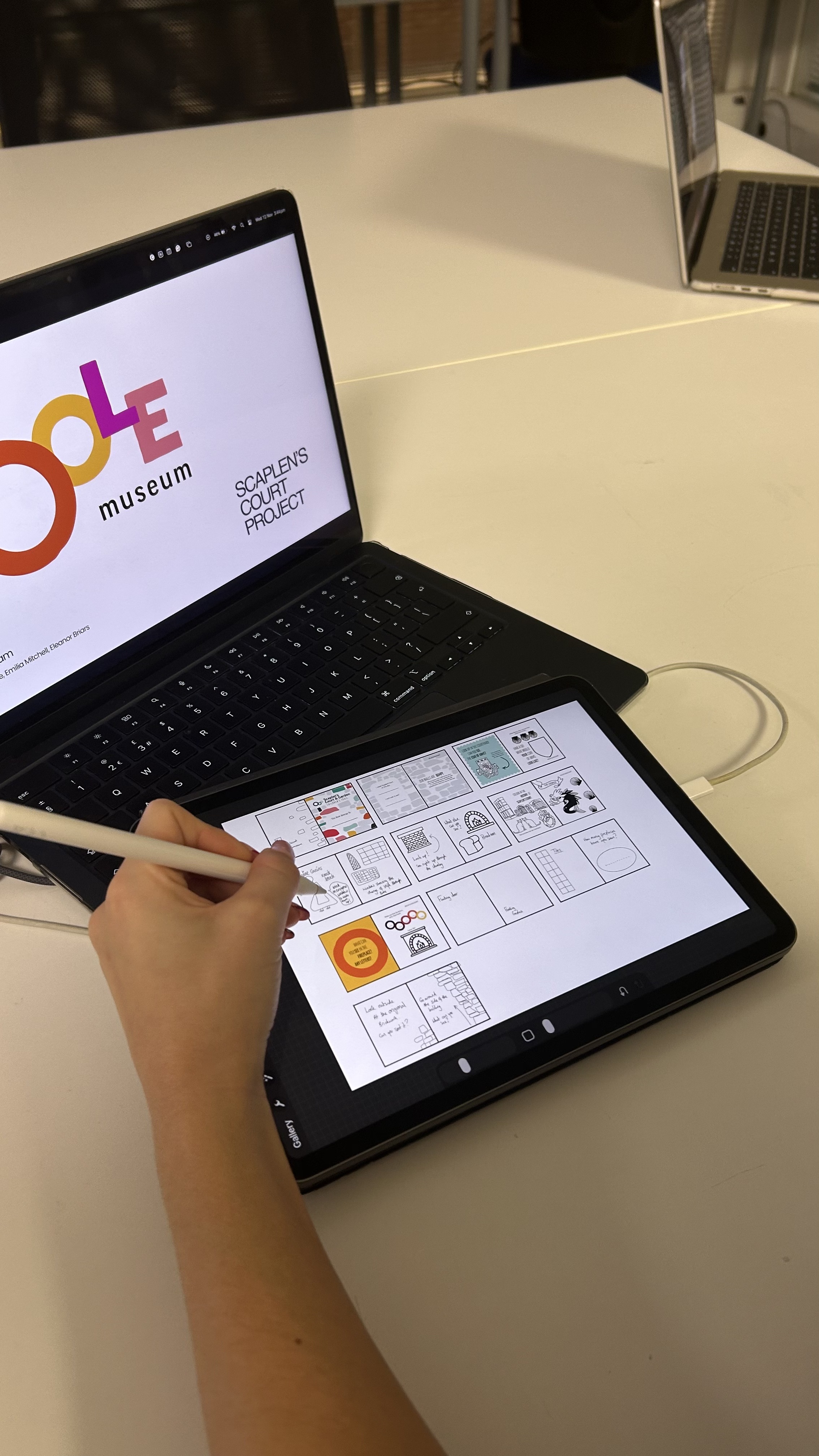 Person using stylus on tablet displaying grid layout whilst laptop shows Ode Museum website with colourful circular logo on white desk.