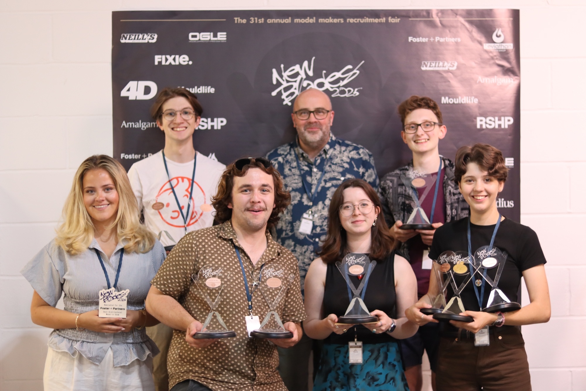 Six BA (Hons) Modelmaking students and course leader P Johnson holding glass awards stand in two rows before a dark sponsor backdrop with white logos.