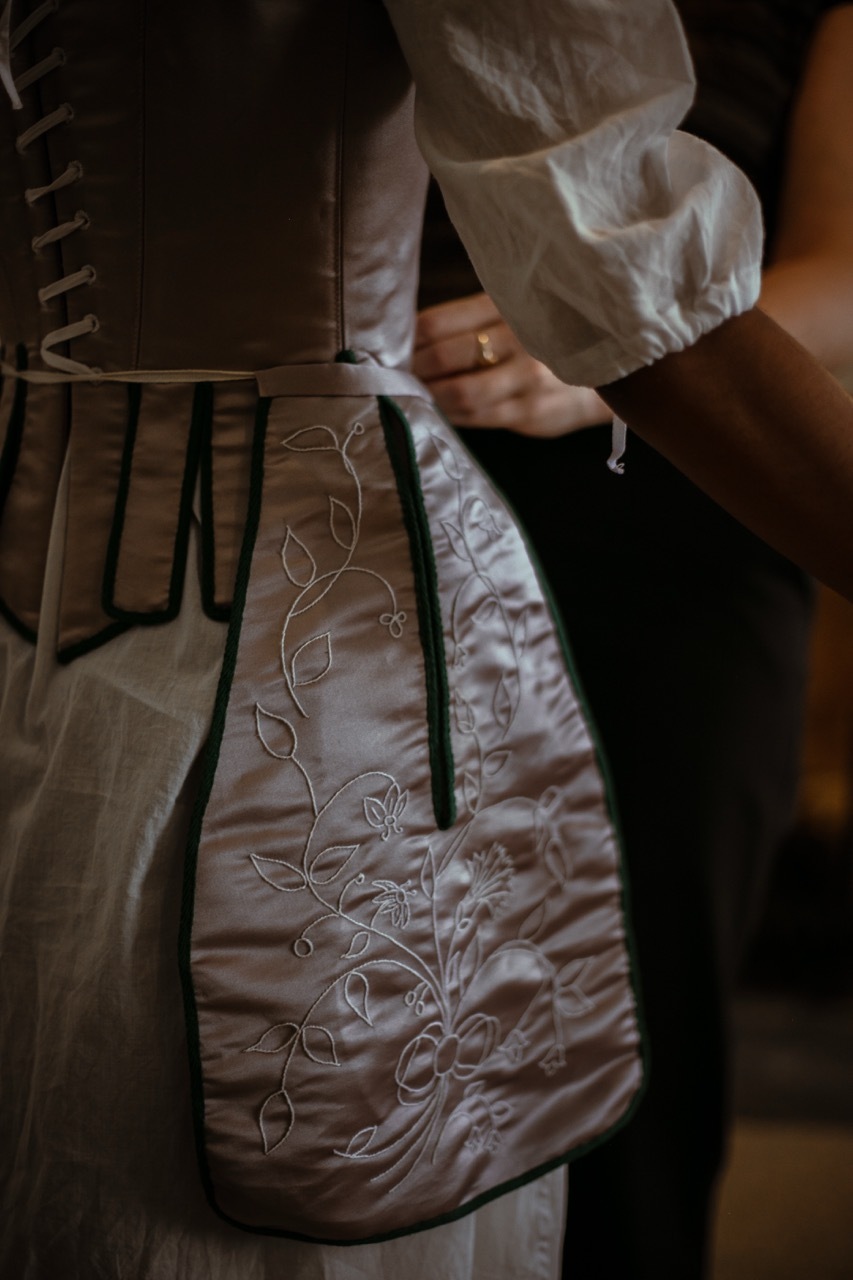 Person in white period costume with embroidered apron featuring floral vine pattern, hands adjusting black skirt or dress.