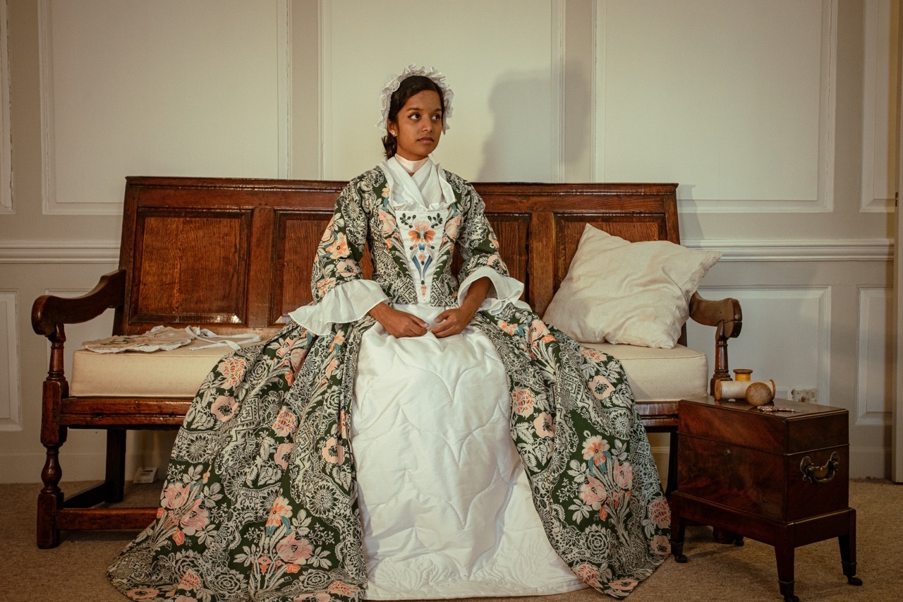 Woman in elaborate 18th-century floral gown sits on wooden daybed in panelled room with white pillows and papers scattered nearby.