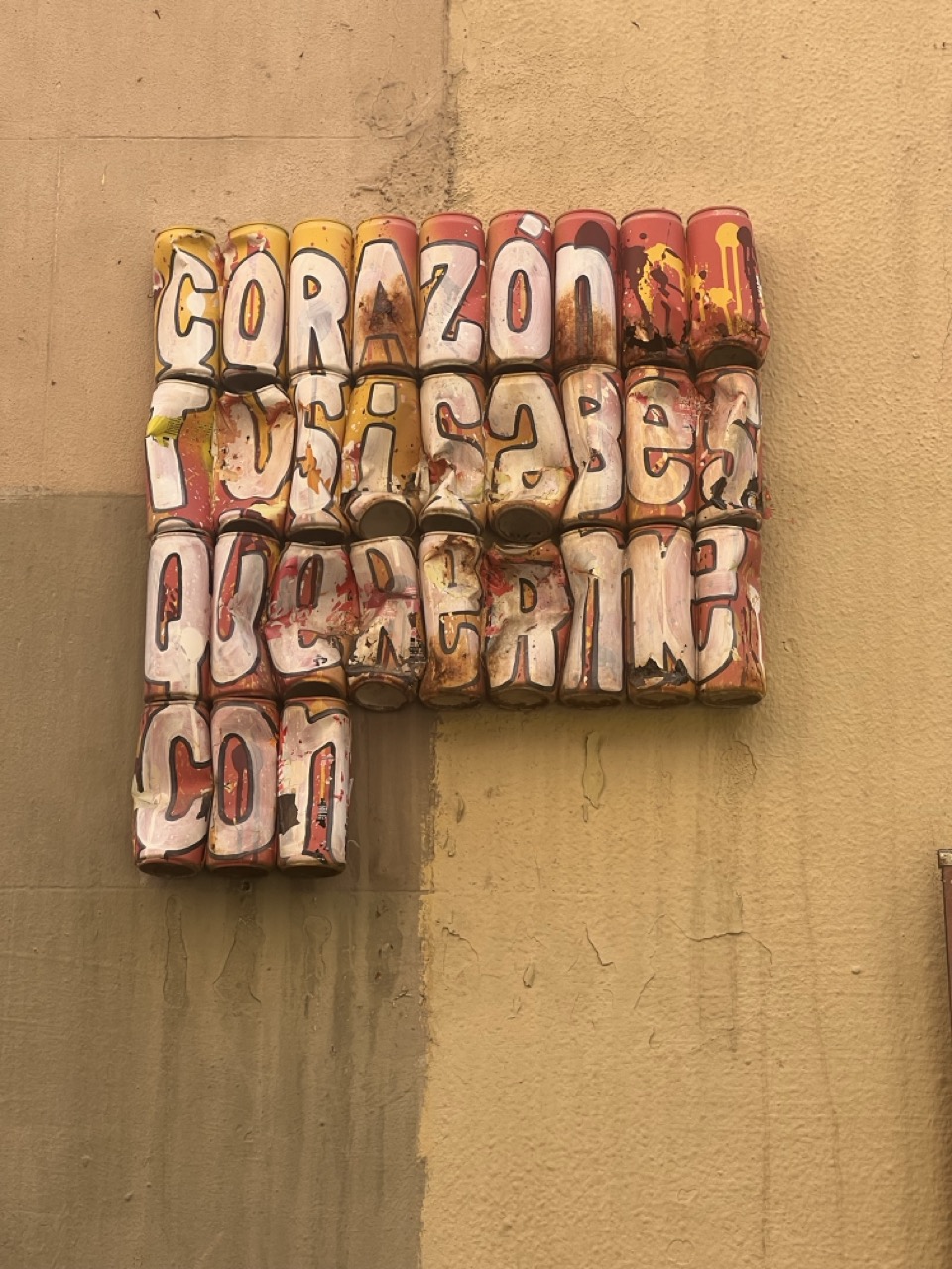 Rows of painted cans in red and yellow with white, funky font letters on the front, earthy coloured wall background.
