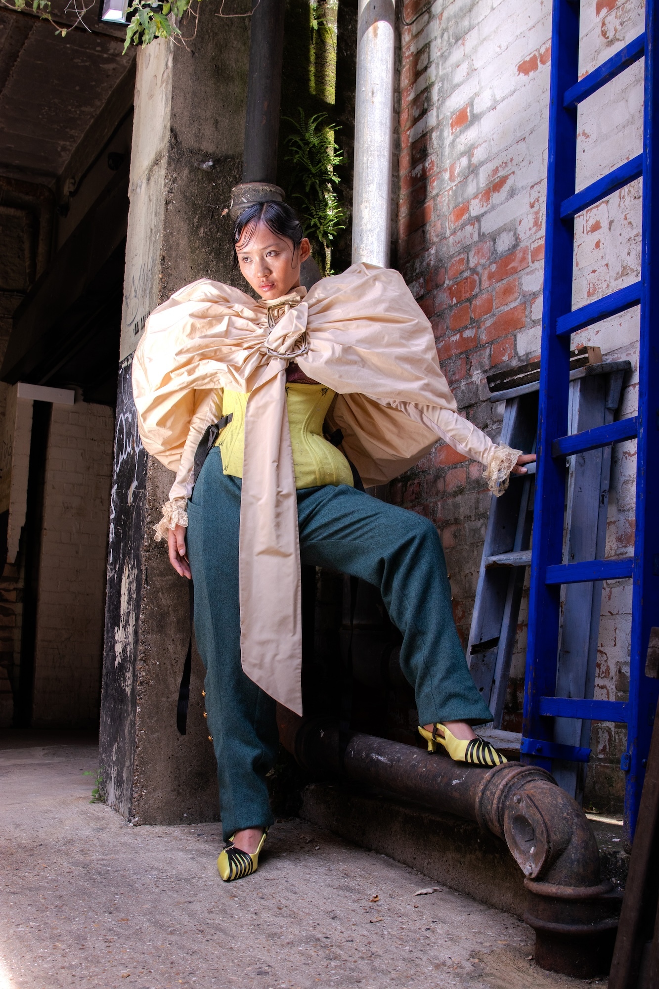 Model wearing a yellow, irregular-shaped top, a pair of dark blue trousers and a pale gold puffy coat/cape, posing with one leg raised on a pipe, next to a blue ladder.