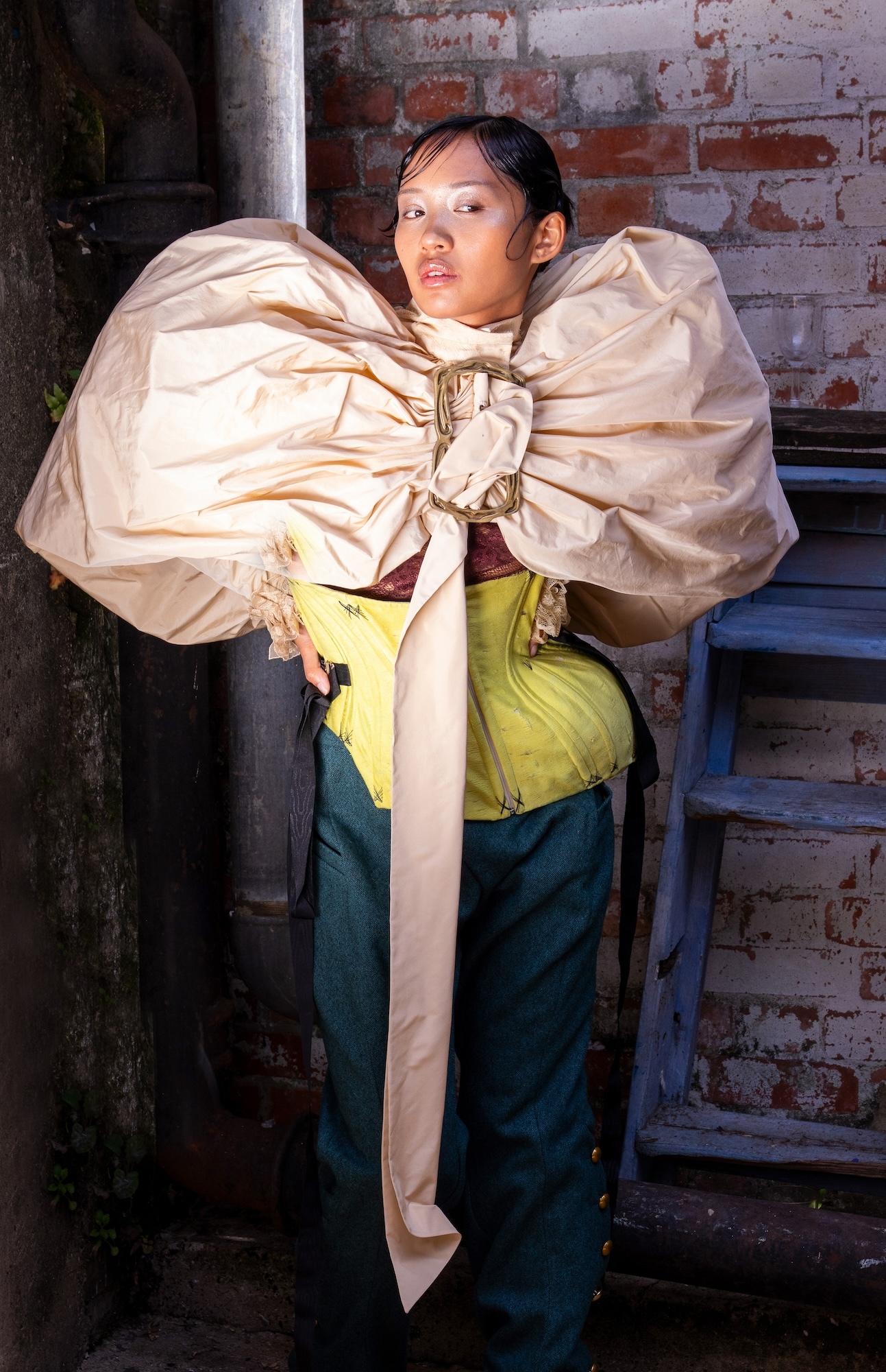 Model posing while wearing a yellow, irregular-shaped top, a pair of dark blue trousers and a pale gold puffy coat/cape.