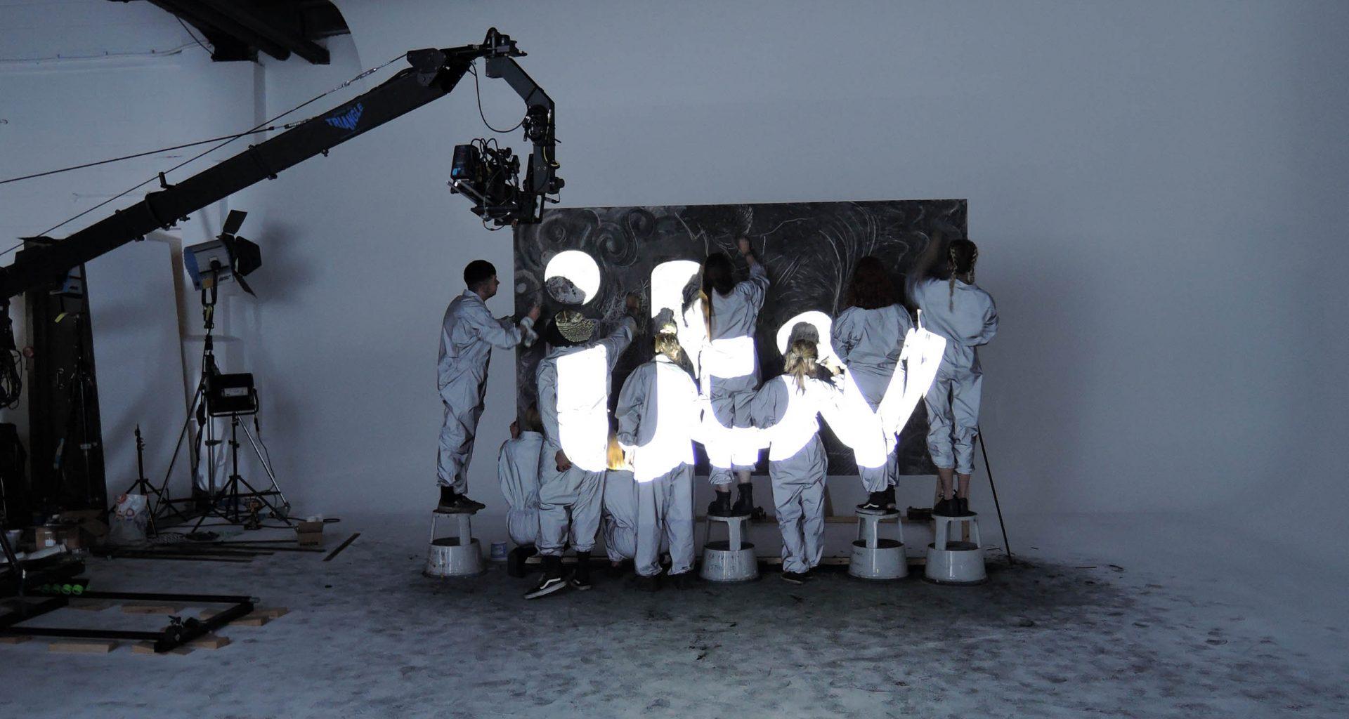 Eight students wearing white uniforms stand in a dark studio space with their backs to the camera working on a painting. The ITV logo is projected onto them.