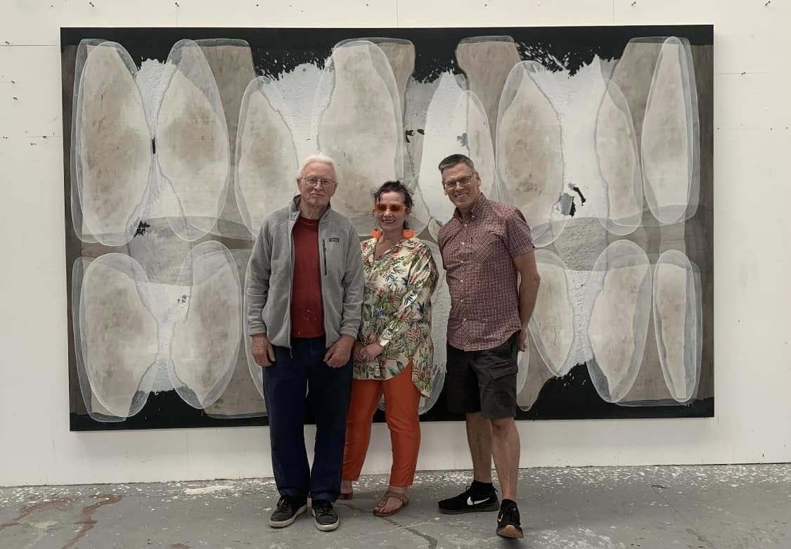Artist I. McKeever with V M McClean and Professor P Gough posing for a photo in front of a large, grey and black abstract painting, displayed on a studio wall.