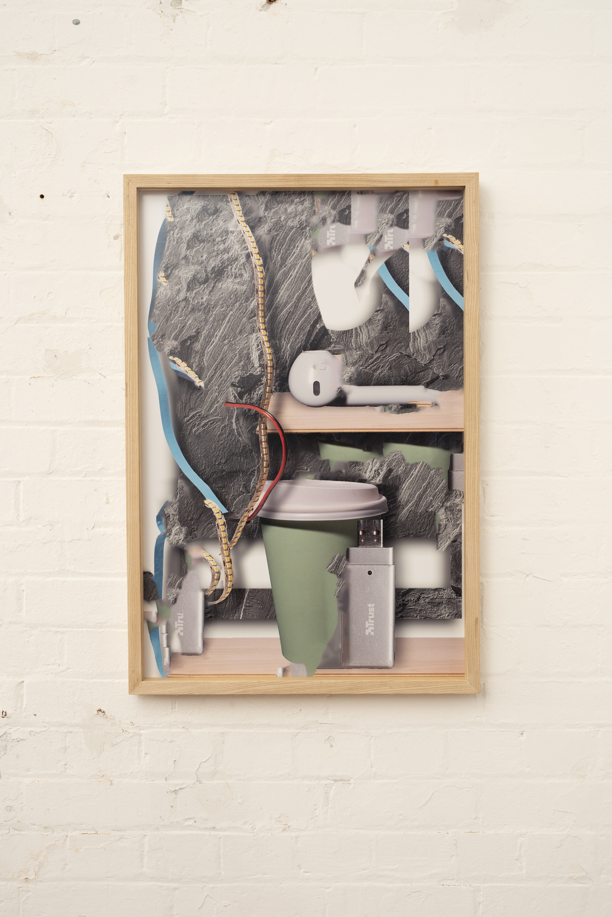 Abstract photographic image of various objects including a USB stick, an earphone and a disposable coffee cup, in a frame against a brick wall.