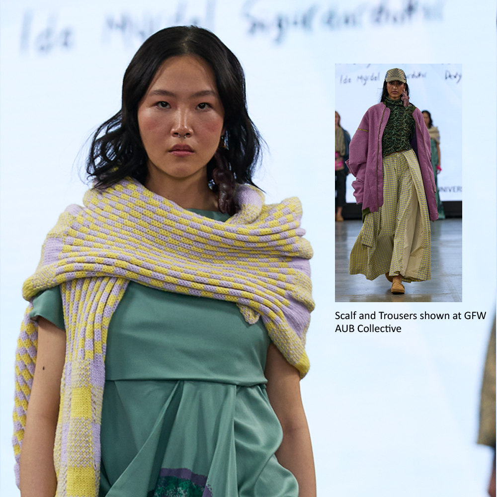 Asian model wearing yellow striped shawl over green dress on runway, with second model in purple jacket and beige trousers visible.