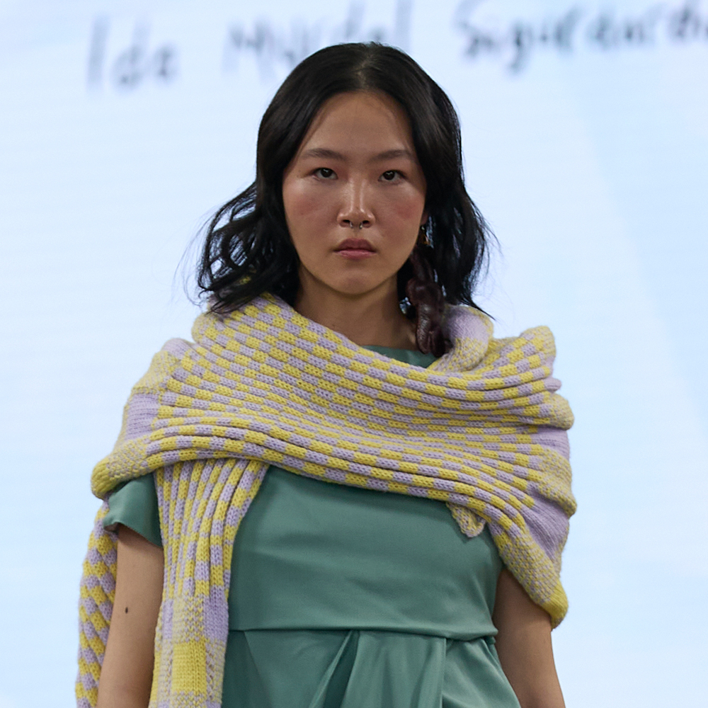 Woman with dark hair wearing mint green dress and yellow-purple striped knitted shawl draped over shoulders against white backdrop.