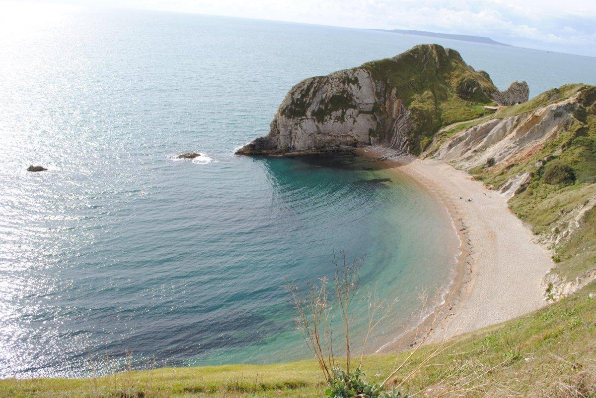 A coved beach on a sunny day, with green, rocky cliffs on the right. The sea is a blue-green and Durdle Door is peaking out behind the cliff.