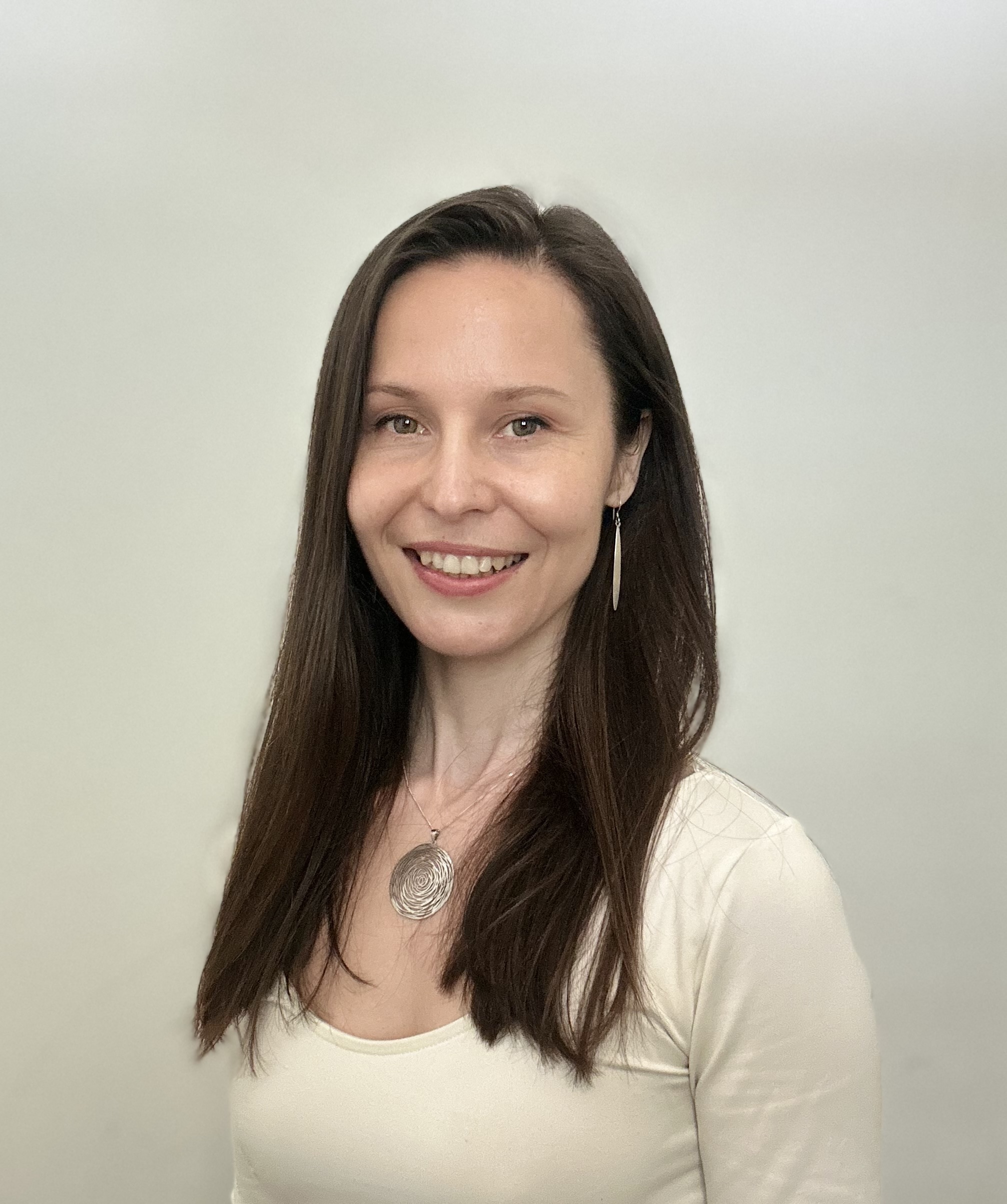 Woman with long brown hair wearing cream top and silver necklace, smiling at camera against light grey background.
