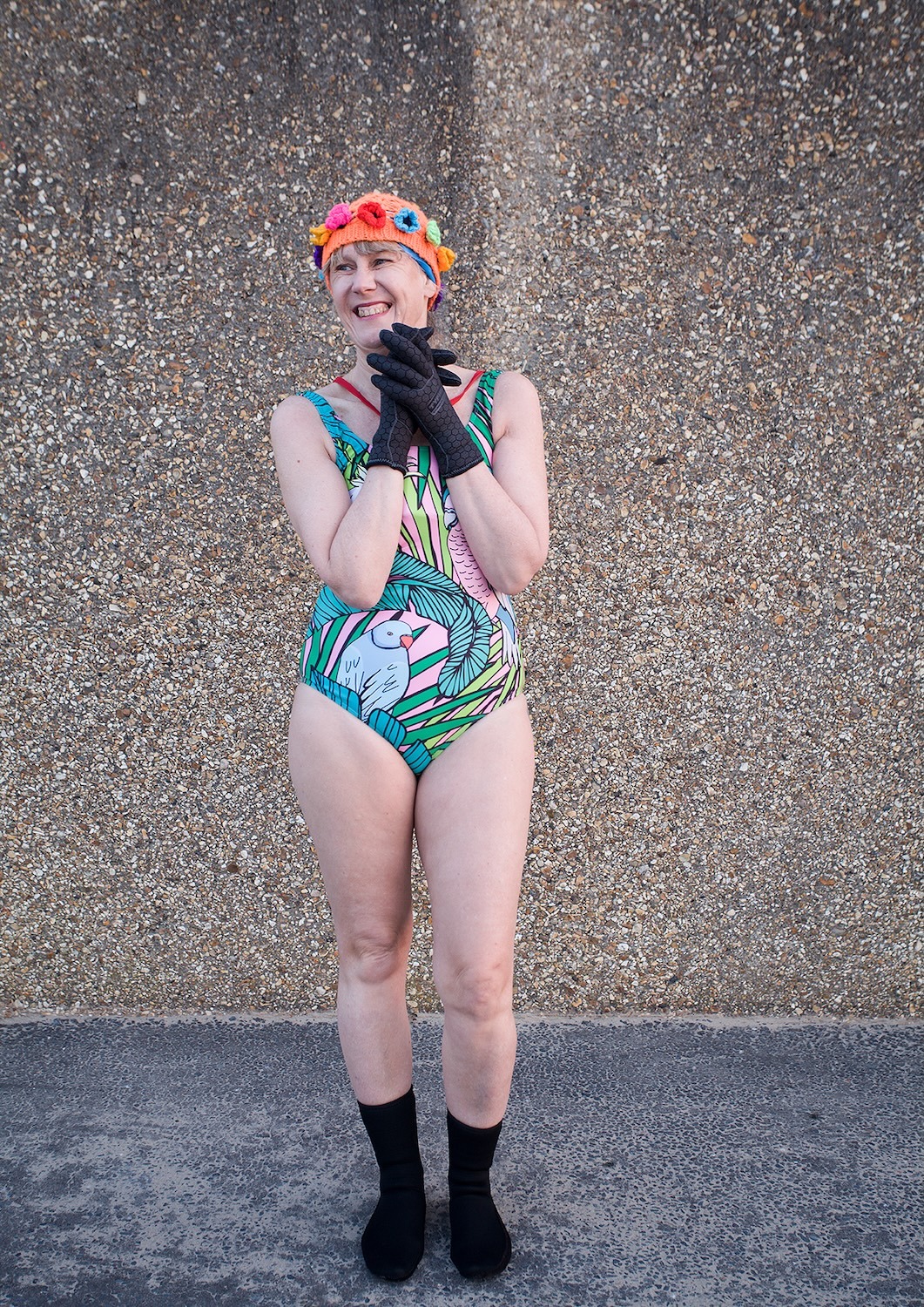 Woman in colourful swimming costume and orange swimming cap standing against pebbled wall, wearing black gloves and socks.