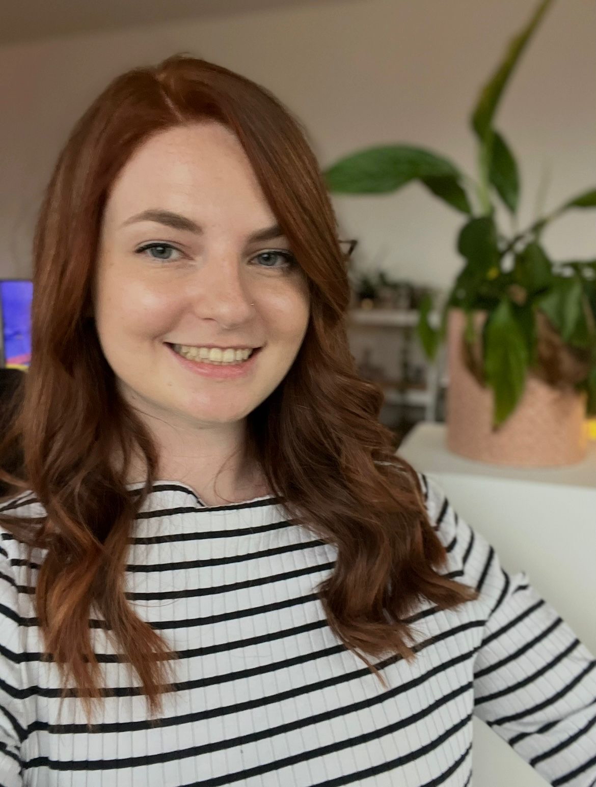 Selfie of Emma McEvoy, with auburn hair and a striped t-shirt, smiling, plant in background.