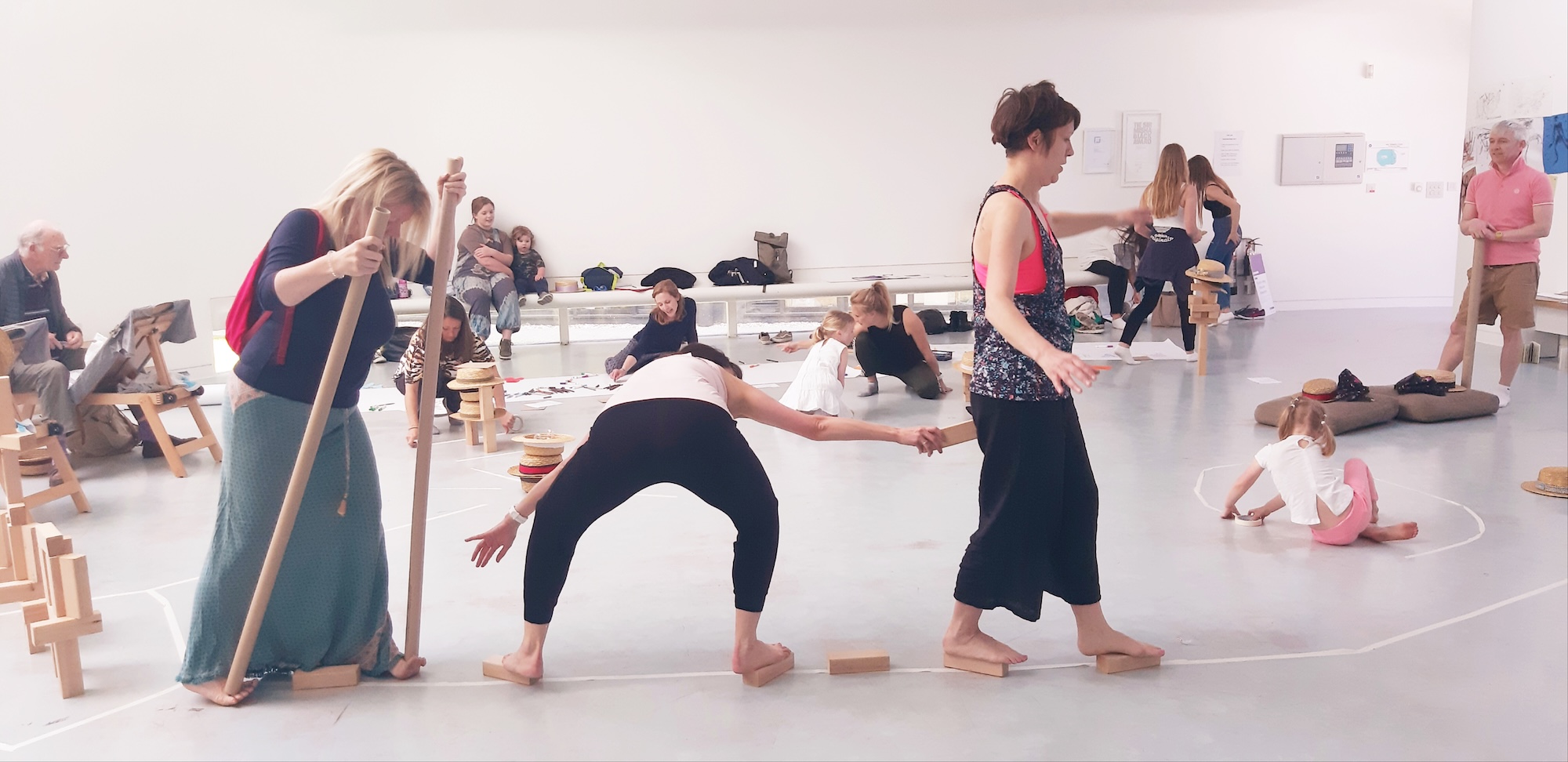Adults practise a group movement class in a bright studio, with rolled mats and wooden blocks scattered across the floor.