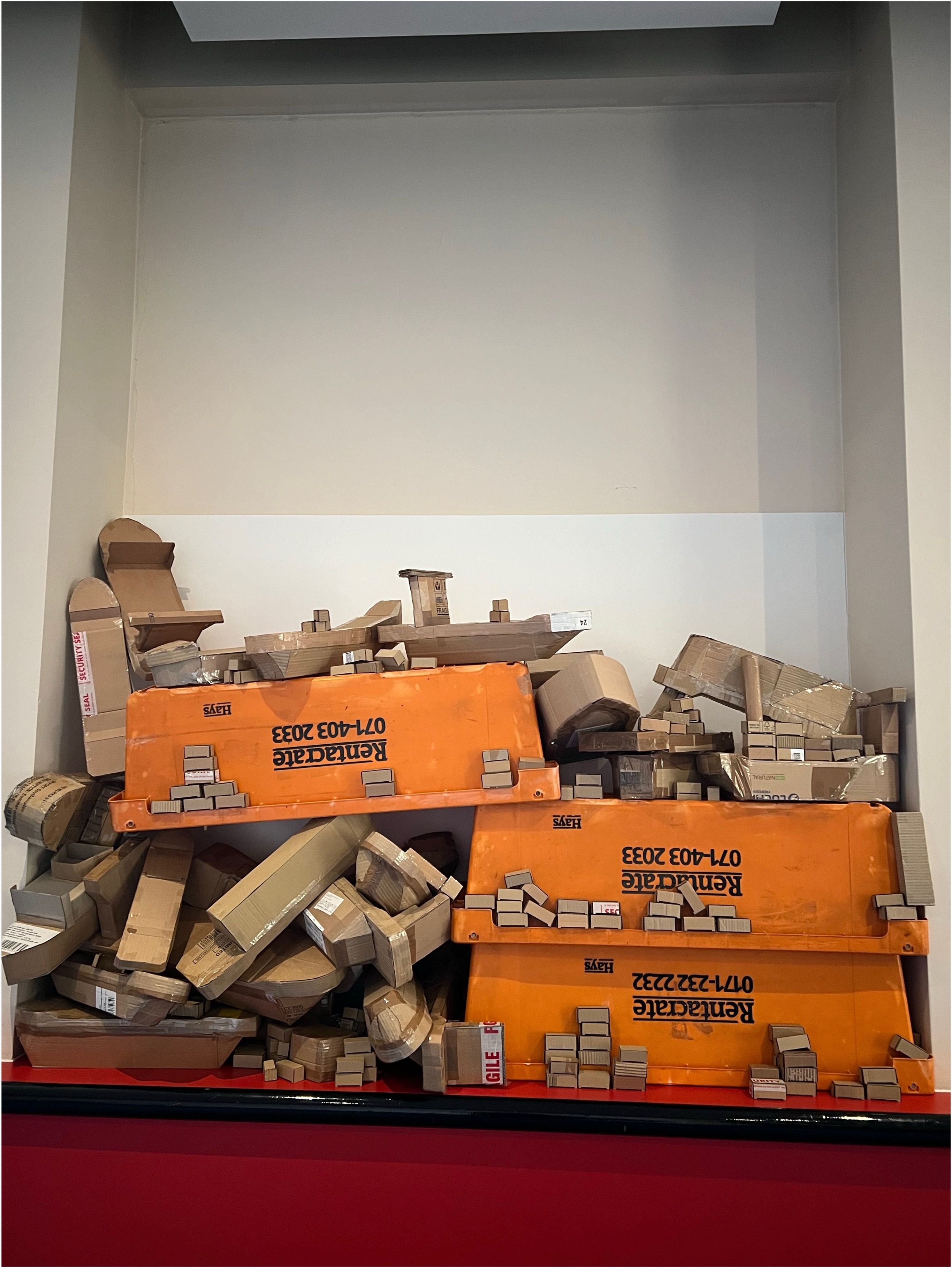 White storage alcove filled with stacked orange Colgate boxes and scattered brown cardboard packages on red surface.