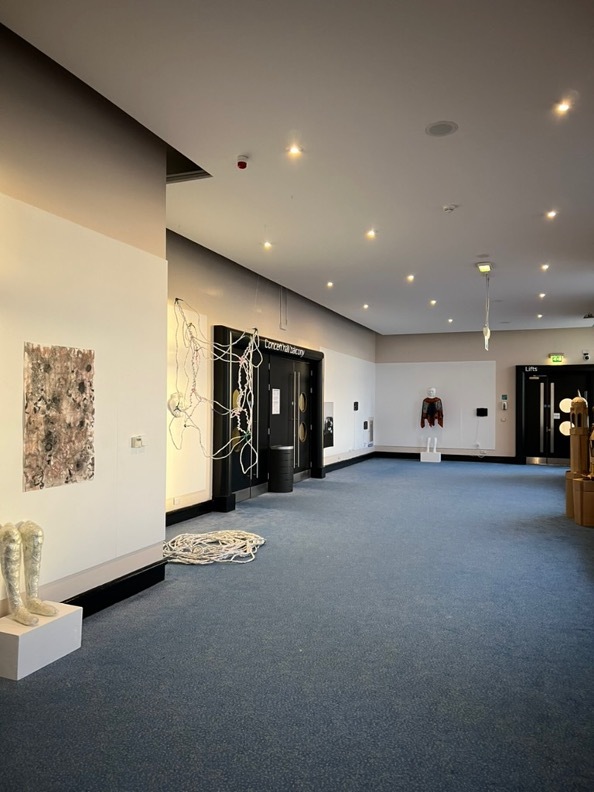 Modern gallery interior with white walls, grey carpet, recessed lighting, and contemporary artworks including sculptures and wall pieces.