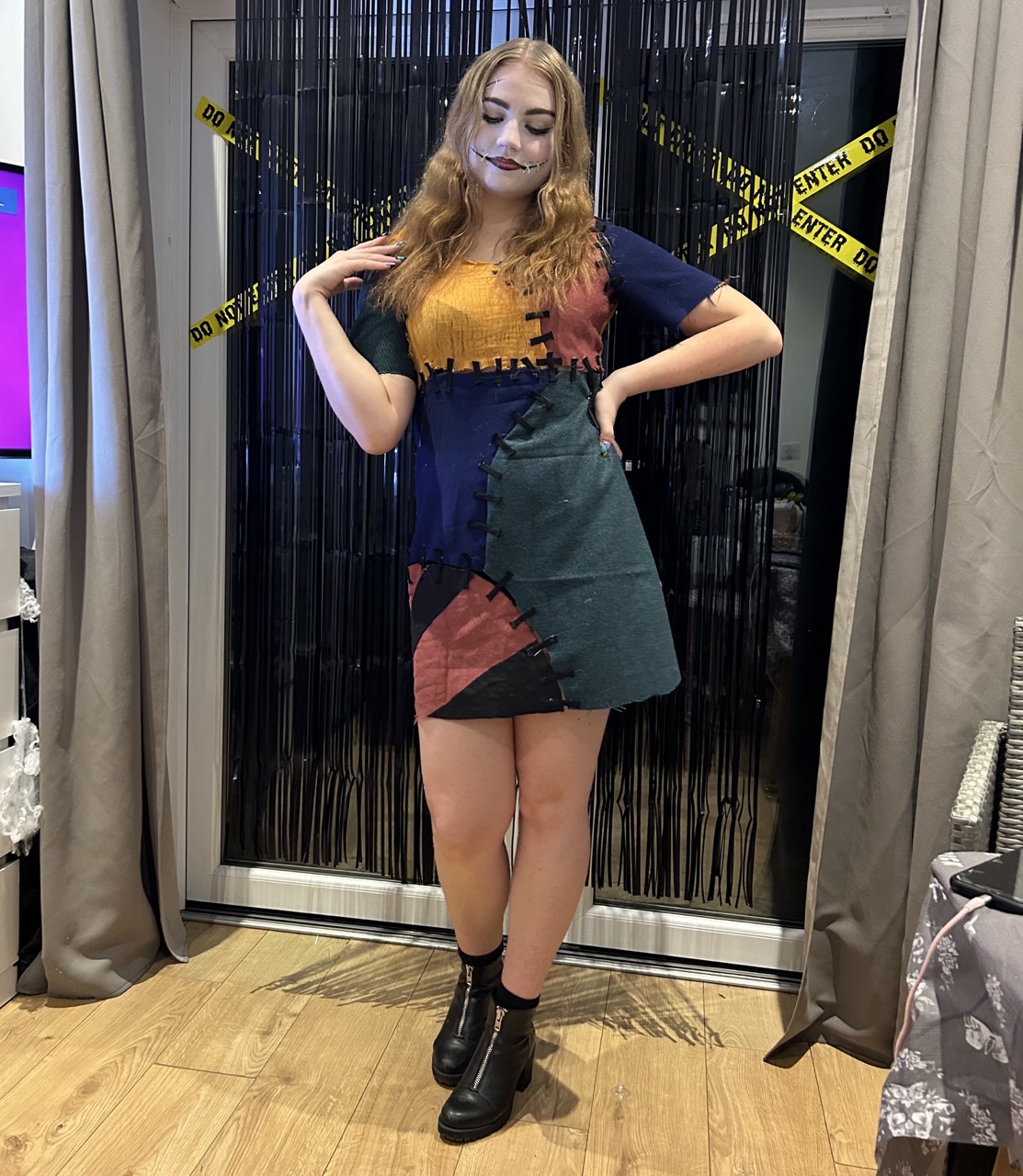 A woman stands posing in front of a black curtain, wearing a multi-coloured patchwork dress featuring blue, green, orange, and red sections. She is wearing black ankle boots and has long, wavy hair styled down. A warning tape is displayed in the background.