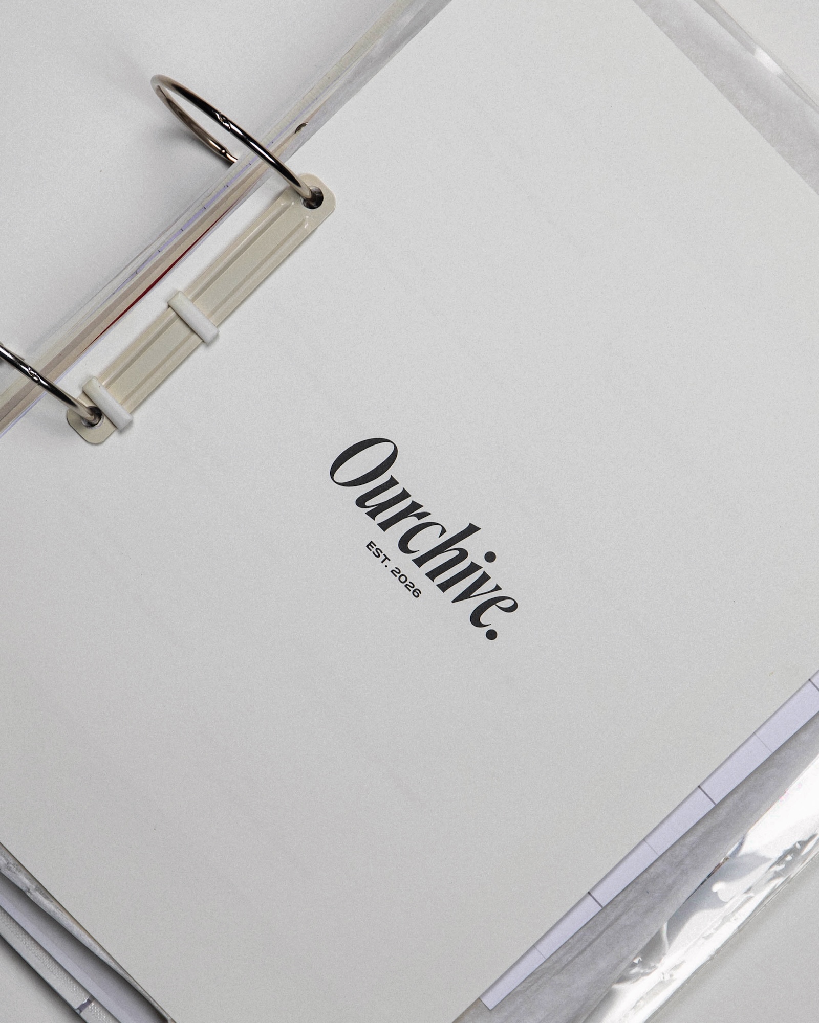 A close-up of a white binder displaying the title 'Ourchive.' with a small subtitle indicating 'EST. 2020'. The binder is secured with a metal ring mechanism, with visible loose papers slightly peeking out from the edges.