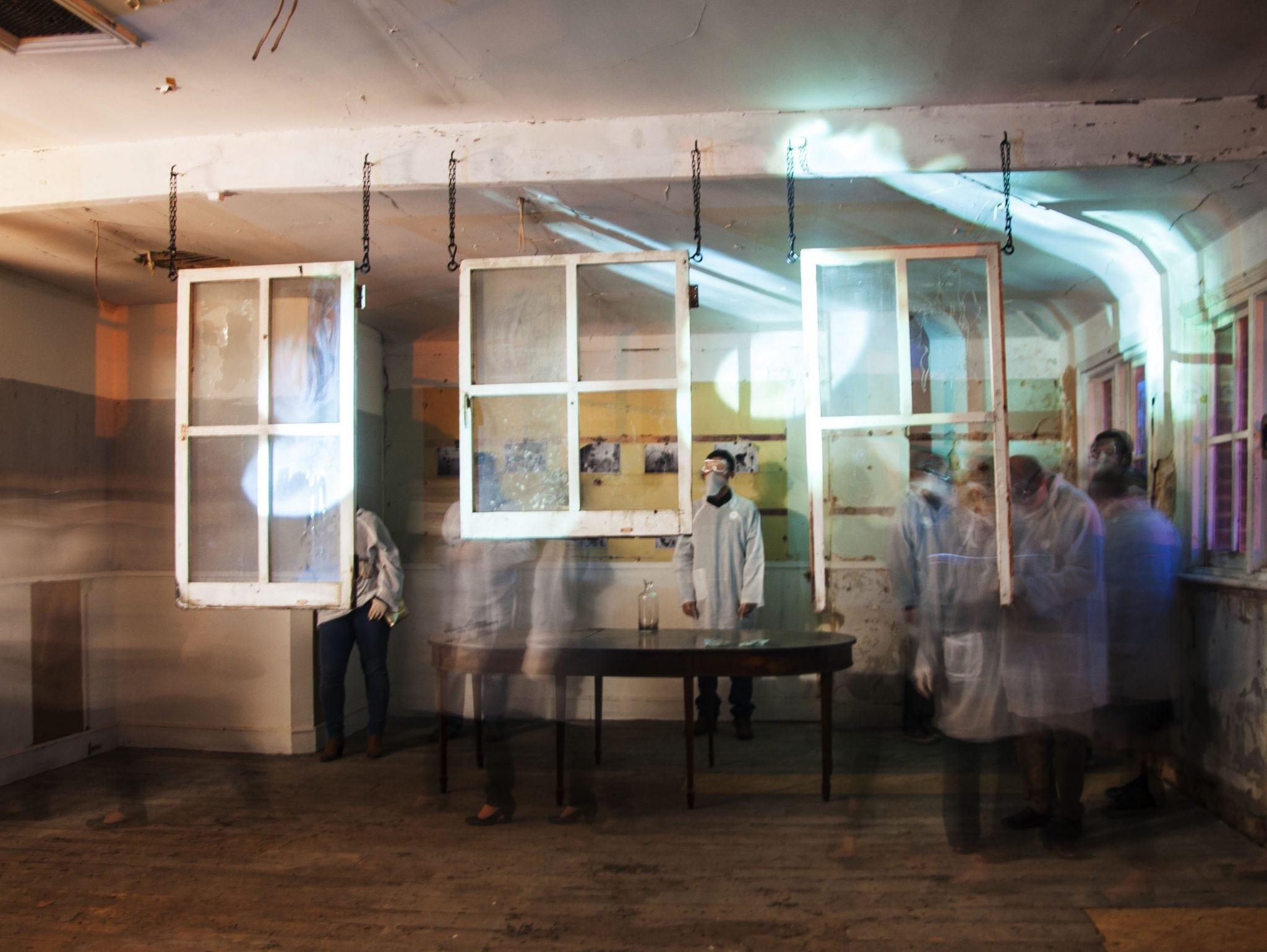 Interior space with suspended white frames and blurred figures moving through green-tinted light, creating ghostly overlapping forms.