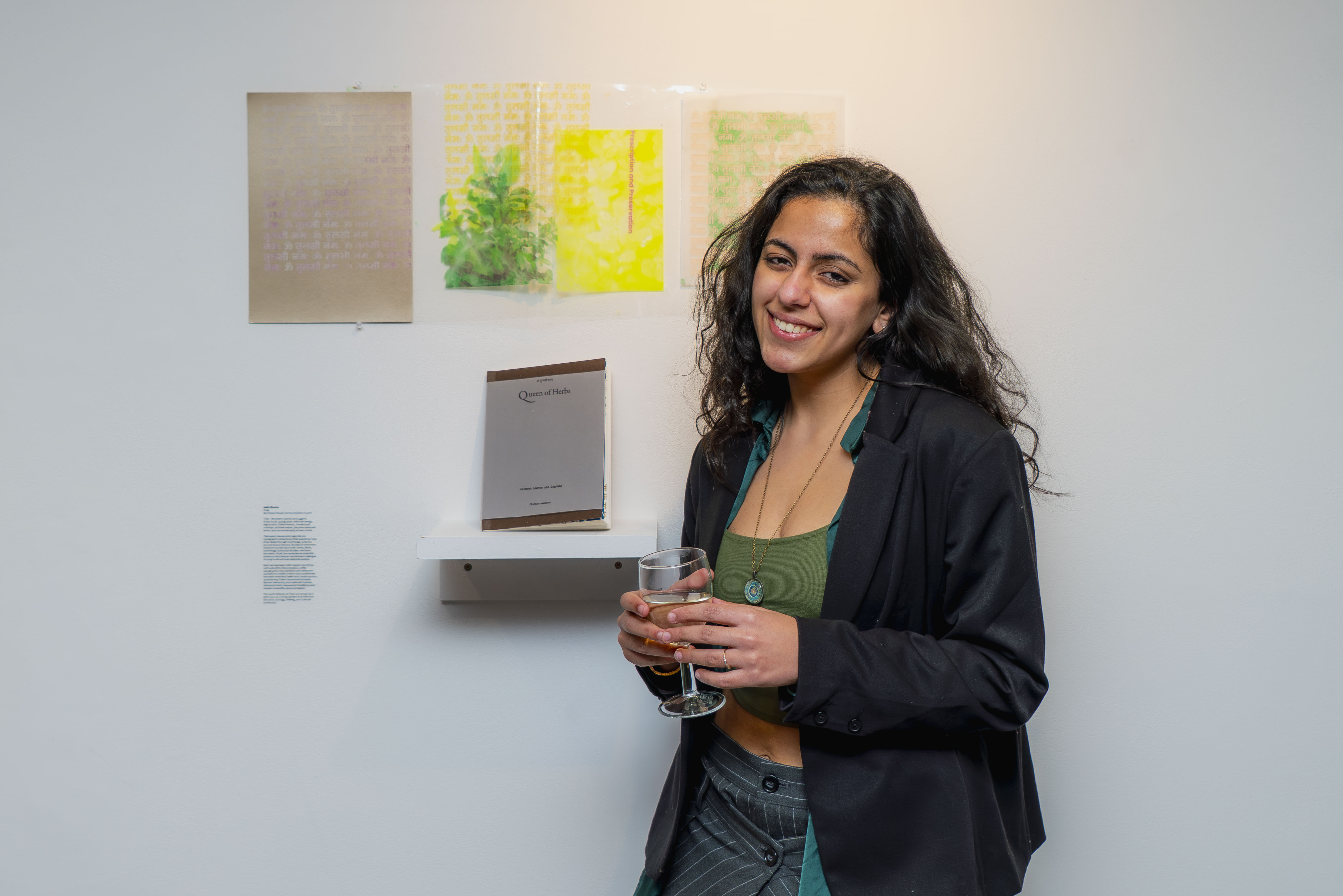Student in black blazer and green top holds wine glass, standing against white wall with framed artworks.