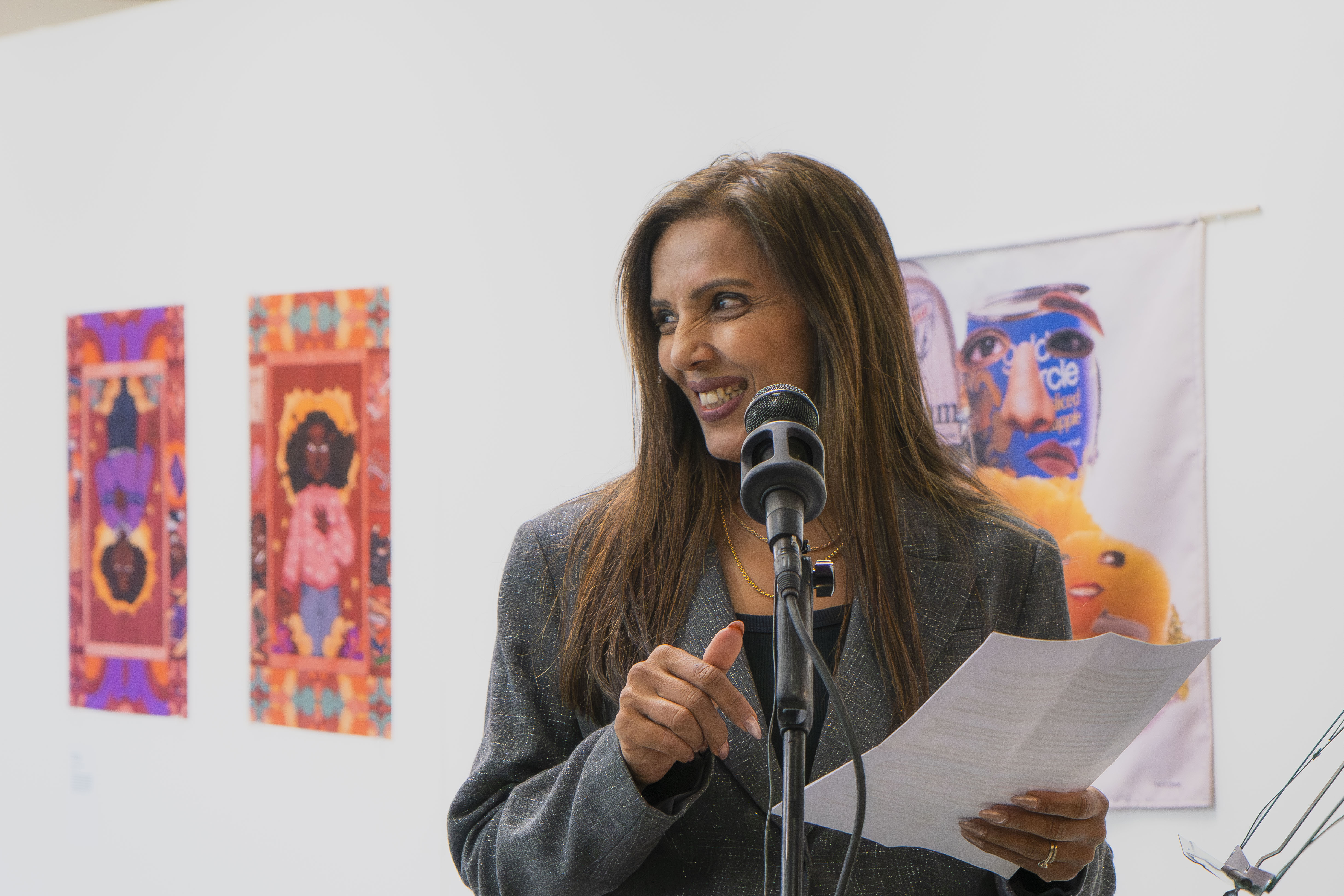 Woman with long brown hair speaking into microphone, holding papers, smiling. Colourful abstract artworks on white wall behind her.