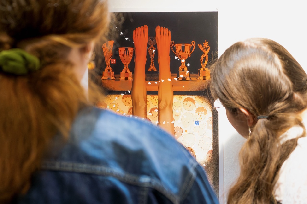 Two students viewing orange abstract artwork with two legs pointing upwards and trophies with illustrated children's faces below, on dark background.