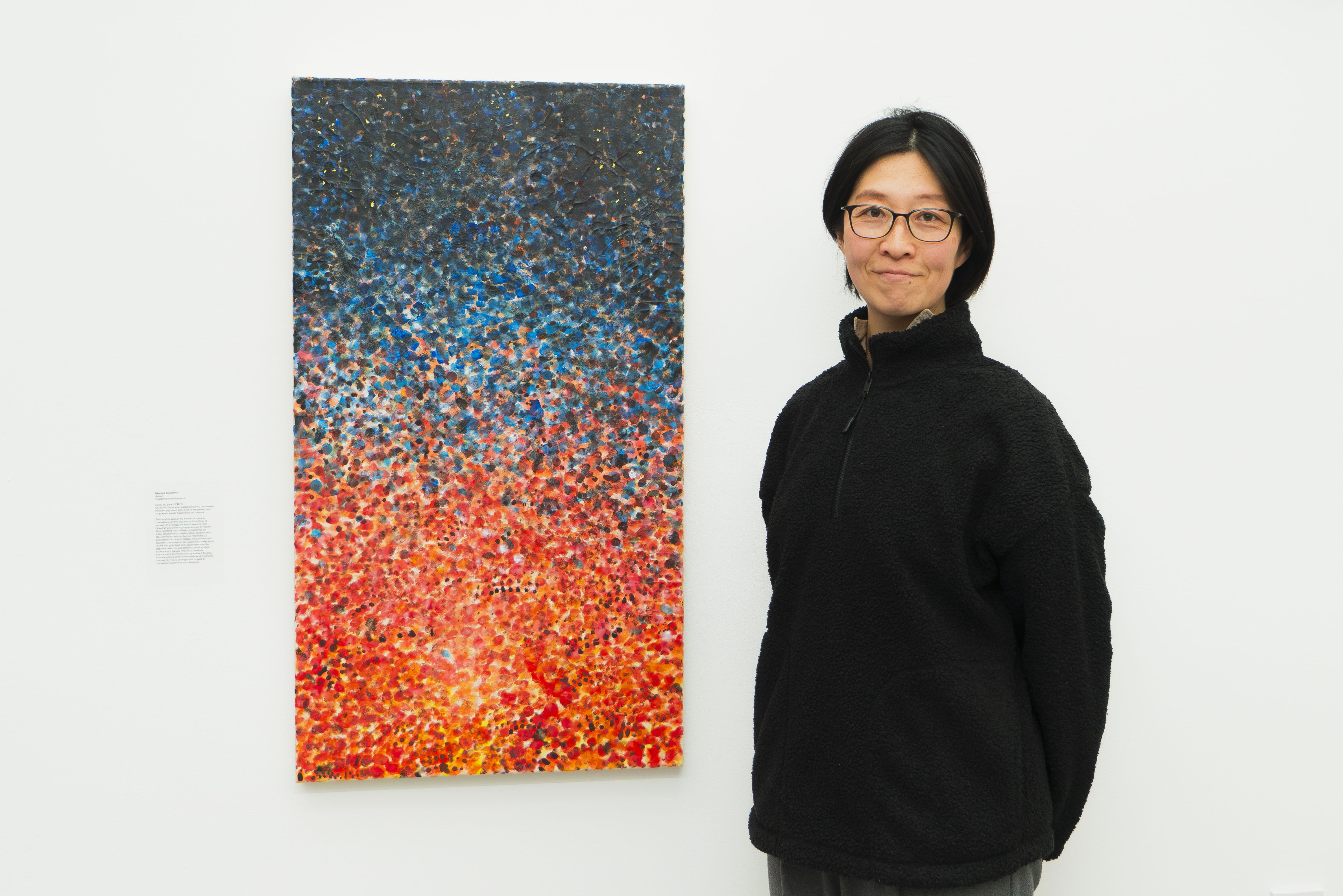 Person in black jumper and glasses standing beside rectangular painting with blue-to-red gradient made of small coloured dots
