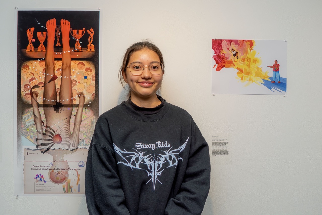 Young woman in dark sweatshirt standing between two artworks on white wall - left piece has orange/brown tones, right has bright colours