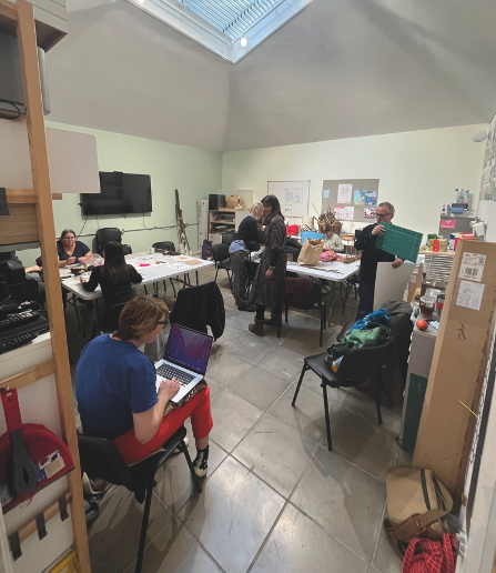 Classroom with students at desks, teacher standing centre, wall-mounted TV, skylight above, tiled floor, storage shelves visible.
