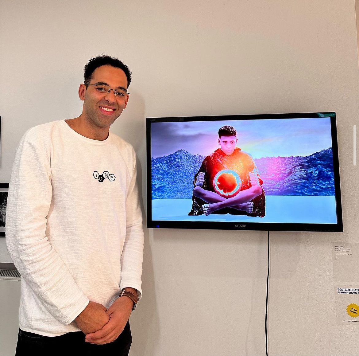 I. Sousa standing next to a monitor at TheGallery, AUB. The monitor screen shows a young person sitting cross-legged with a glowing red sphere in his lap.
