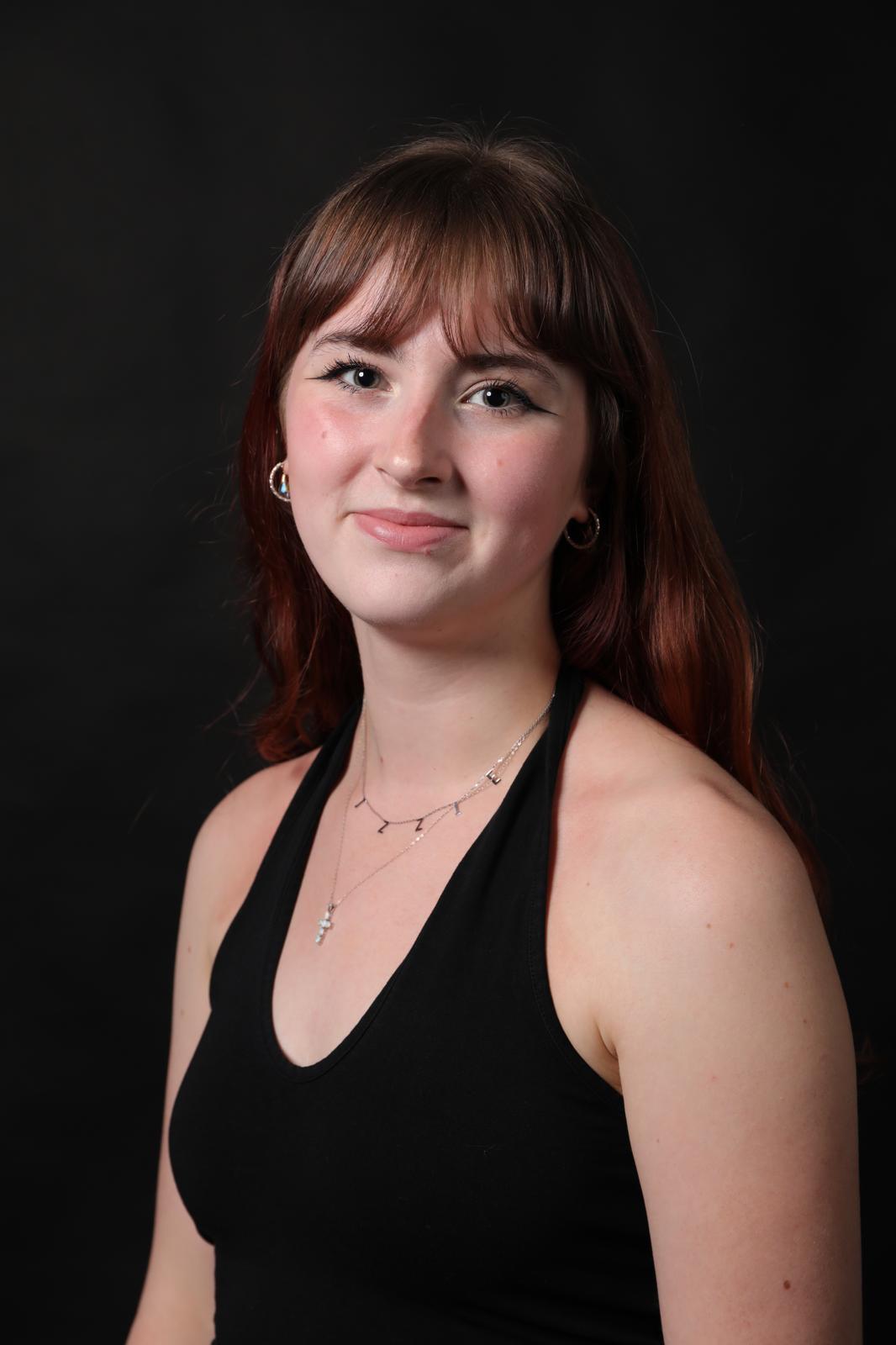 Woman with brown hair and fringe wearing black halter top and delicate silver necklace, smiling against dark background.
