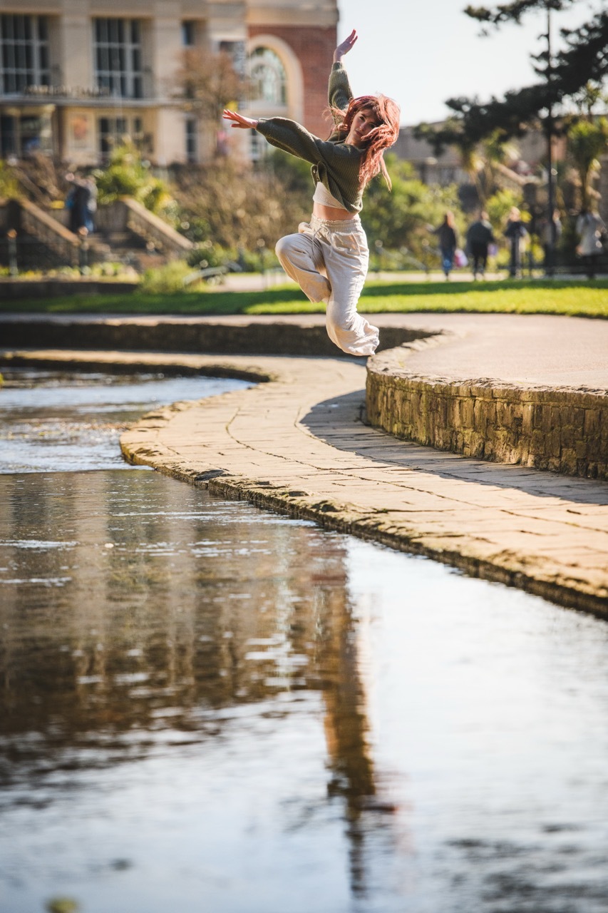 Woman jumping in mid-air with arms outstretched over the river in Bournemouth Gardens, with buildings and trees in background.