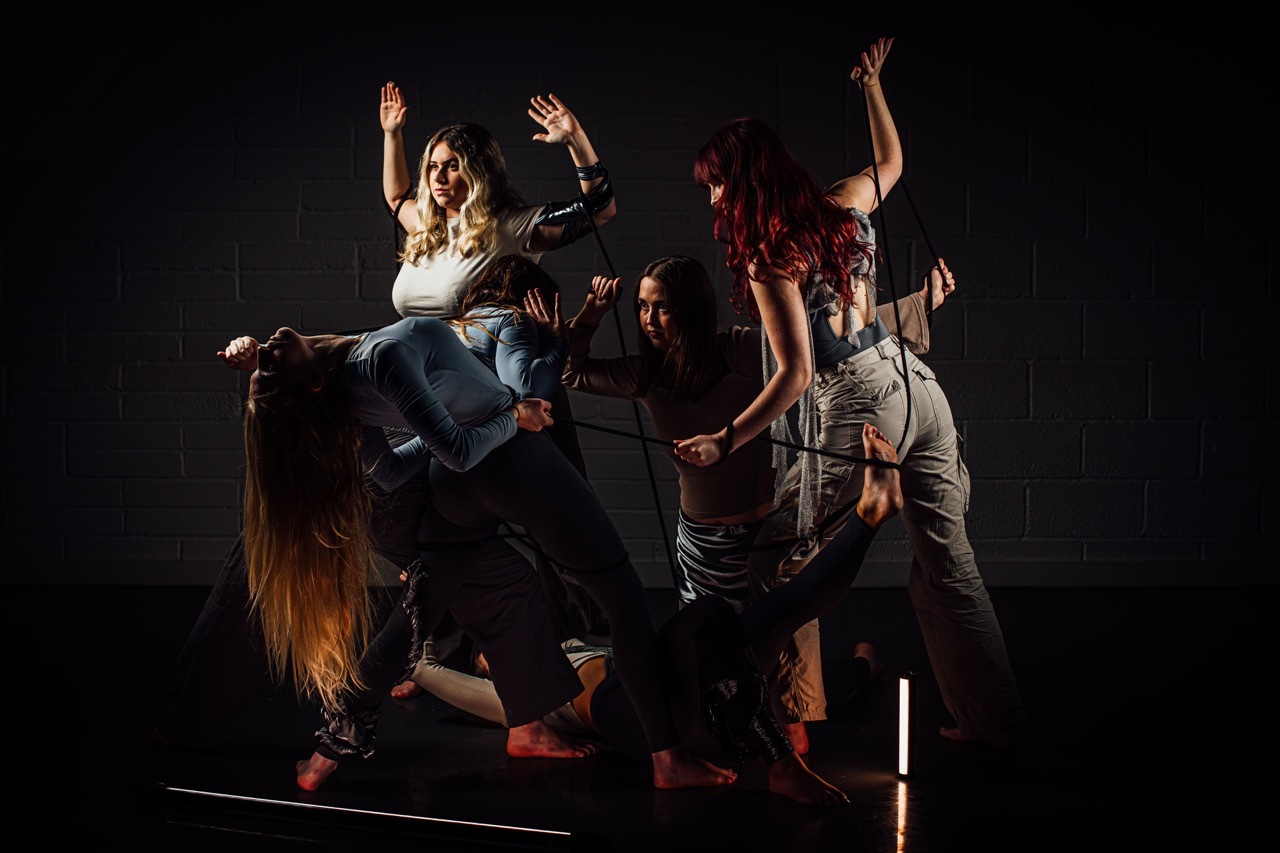 Five performers on dark stage with dramatic lighting, arms raised in dynamic poses, wearing casual clothing in muted colours.