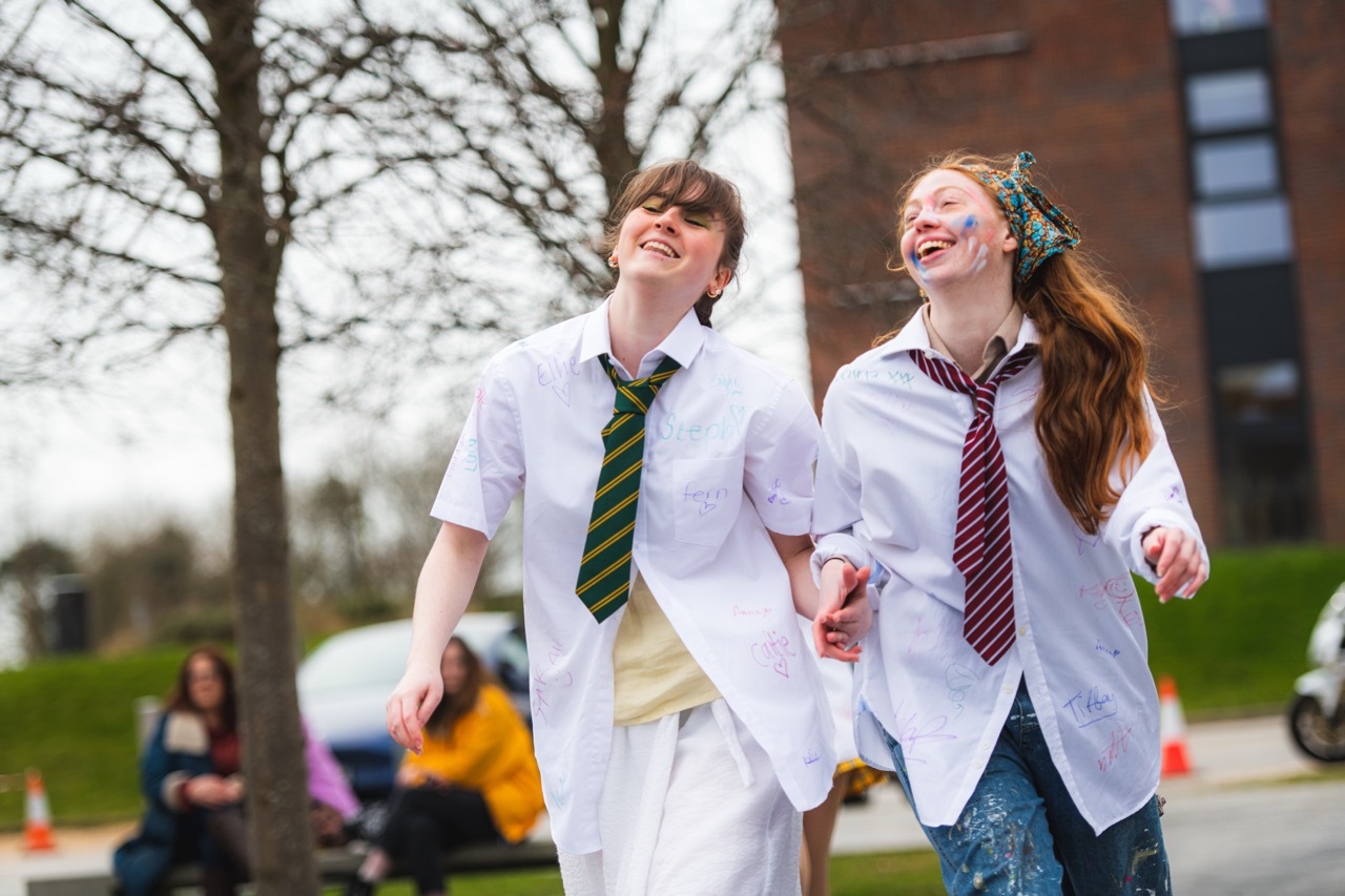 Two teenagers in white shirts and school ties running hand-in-hand across AUB campus, with halls of residence and bare trees behind.