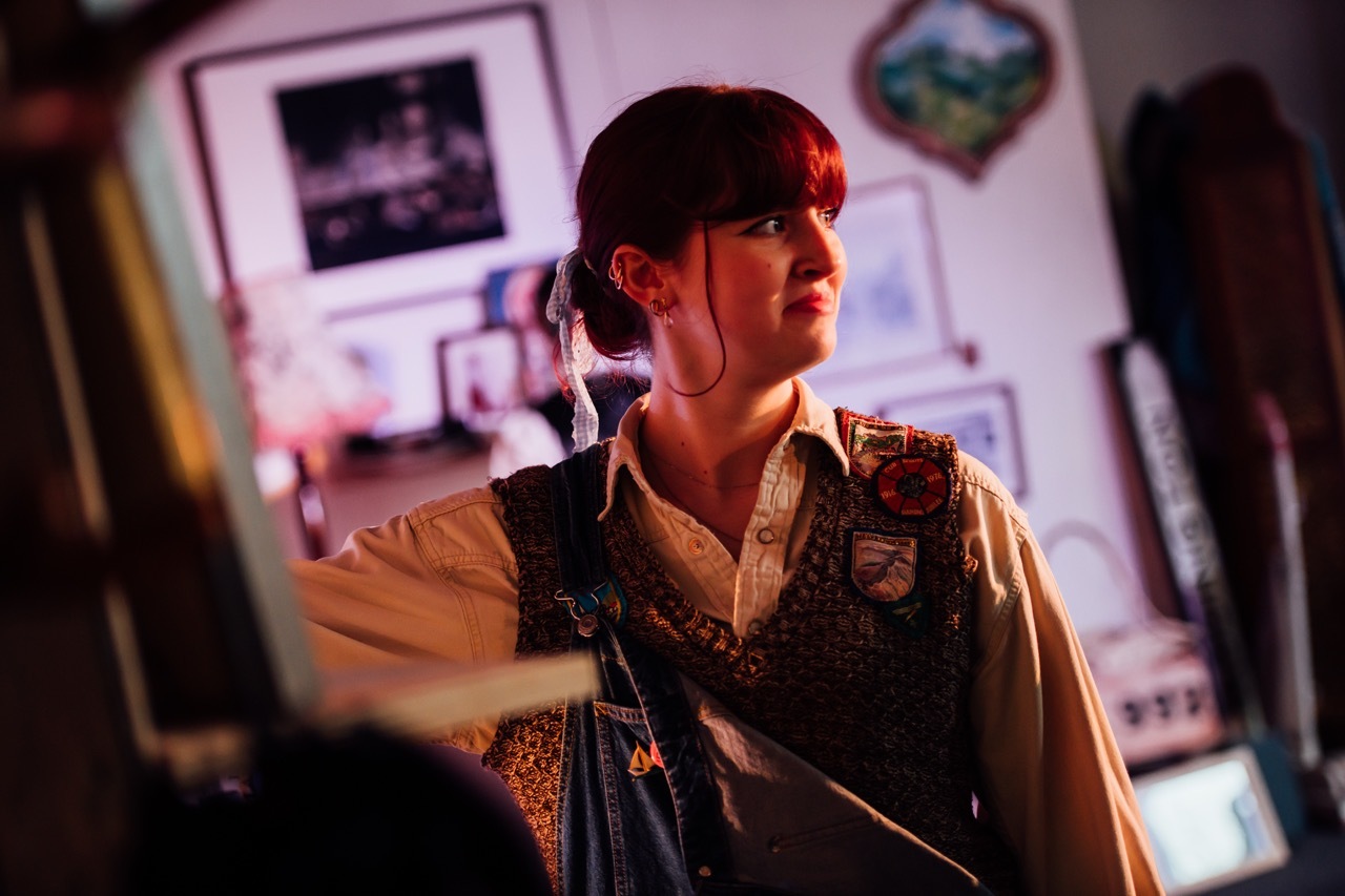 Woman with dark hair in bun wearing patterned green vest over beige shirt, illuminated by red lighting in cluttered room with posters.