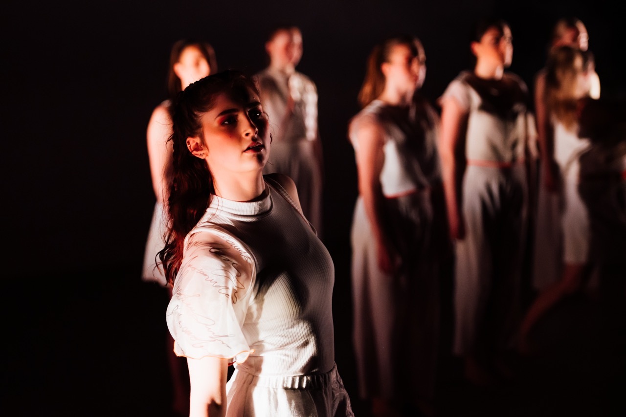 Female performer in white clothing stands in foreground with group of dancers in white garments behind her against dark background.