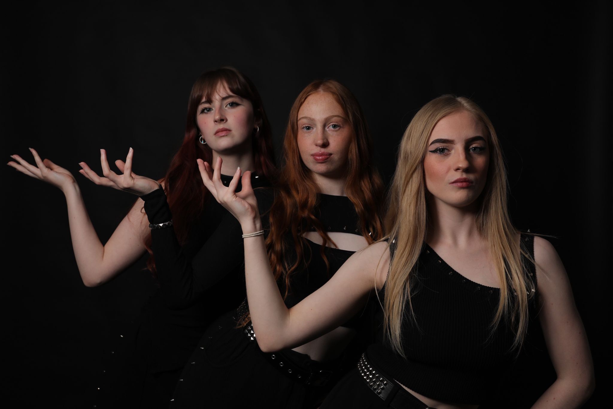 Three women in black clothing against dark background. Left woman has brown hair, hands raised with clawed fingers. Centre woman has long ginger hair. Right woman has blonde hair.