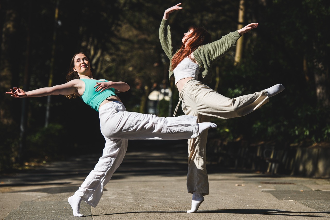Two dancers leaping simultaneously outdoors, one in turquoise top with white trousers, other in green top with beige trousers, trees behind.