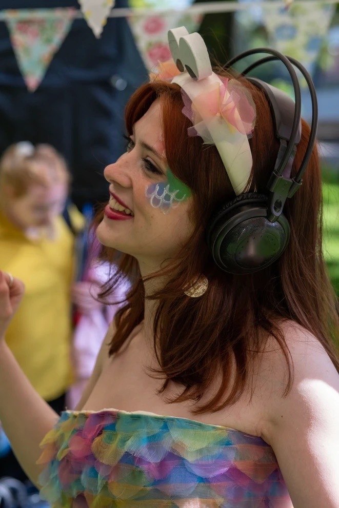 Person wearing colourful dress and make-up with bunny ears and headphones stood outside smiling, blurred bunting and child in the background.