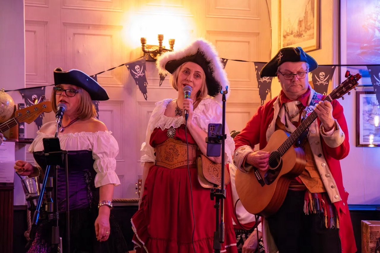 Singers and musicians performing in Pirate costumes in a large room, moody warm and purple lighting.