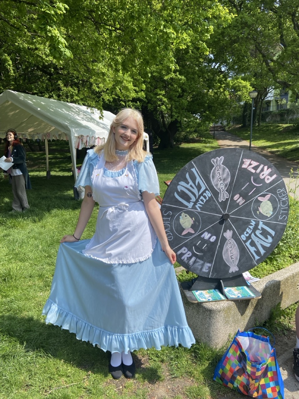 Blonde-haired woman stood outside on the grass dressed as Alice from Alice in Wonderland, stood next to a chalkboard prize spinning wheel, white marquee in the background.