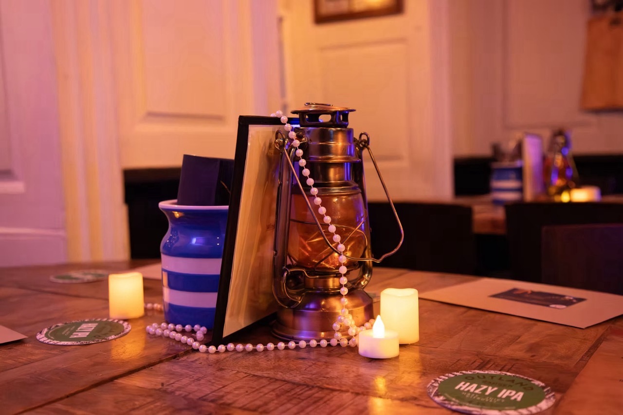 Oil lamp, book with white pearls and LED candle props stacked on a wooden table, moody orange lighting.