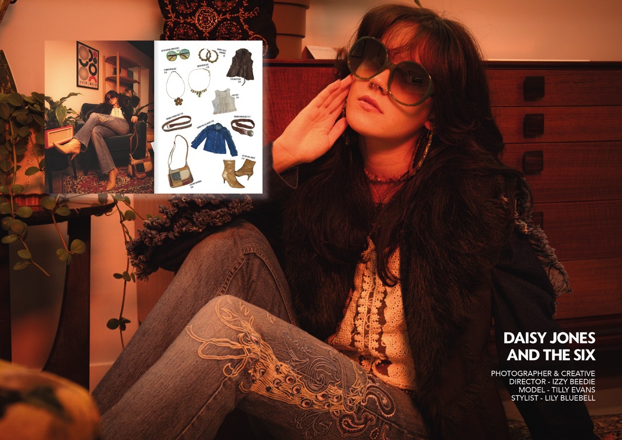 Woman with dark hair and sunglasses sits in orange-lit room with hand to head, surrounded by wall art and posters.