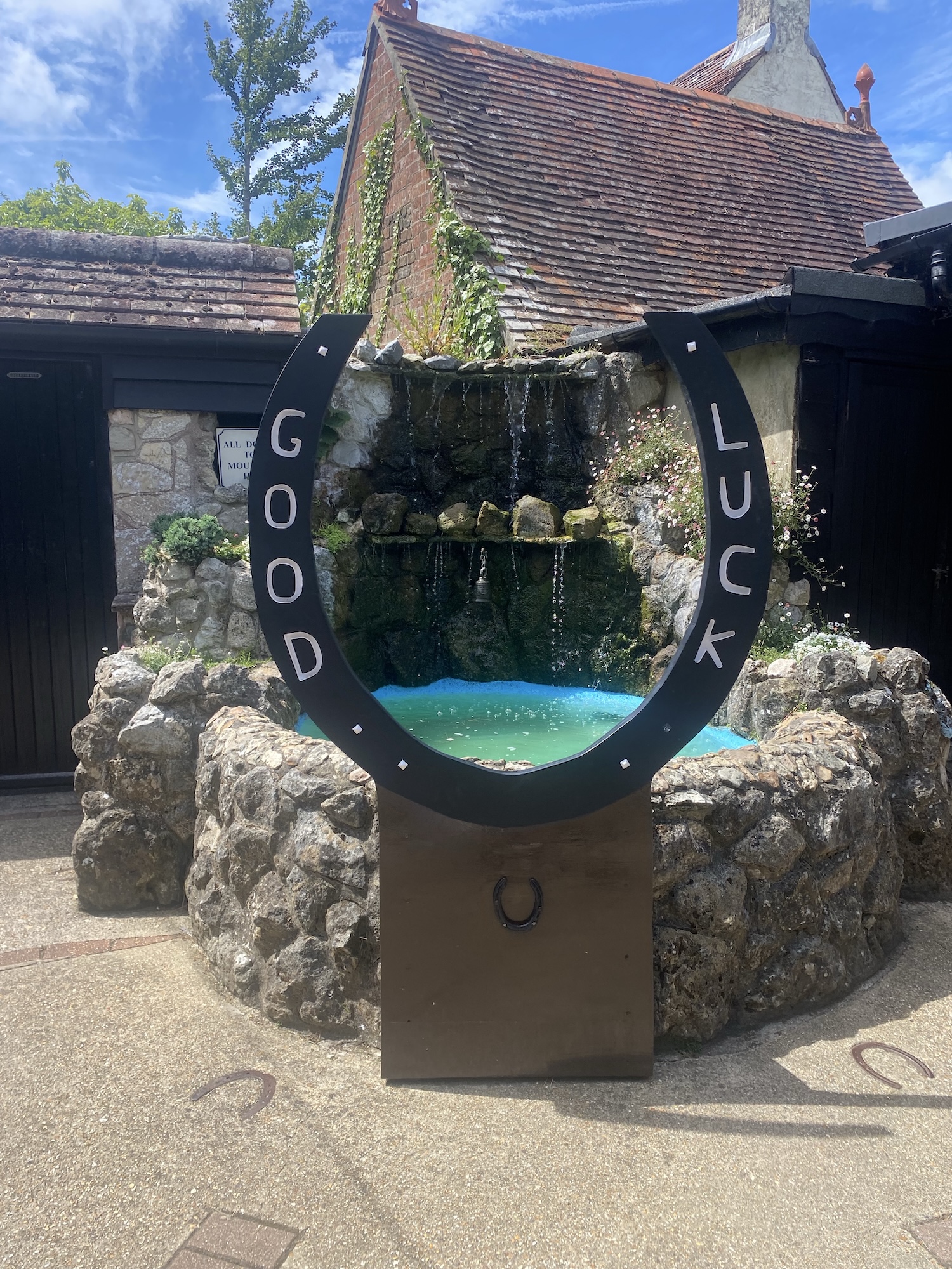 A black horseshoe sculpture marked GOOD LUCK stands in front of a small rock fountain, with rustic buildings and a blue sky in the background.