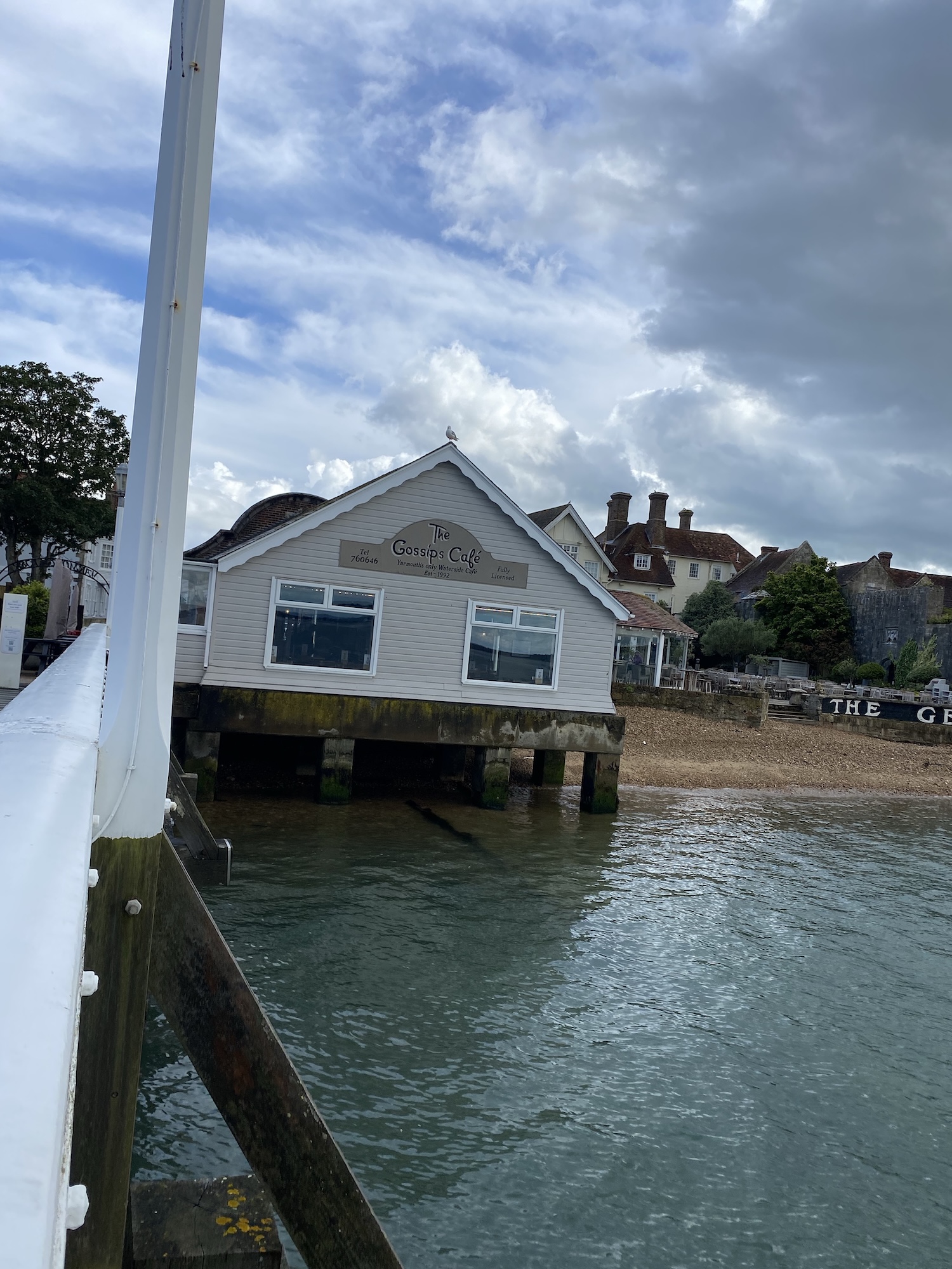 A grey seaside building on stilts beside calm water, viewed from a pier with a white railing under a cloudy sky.