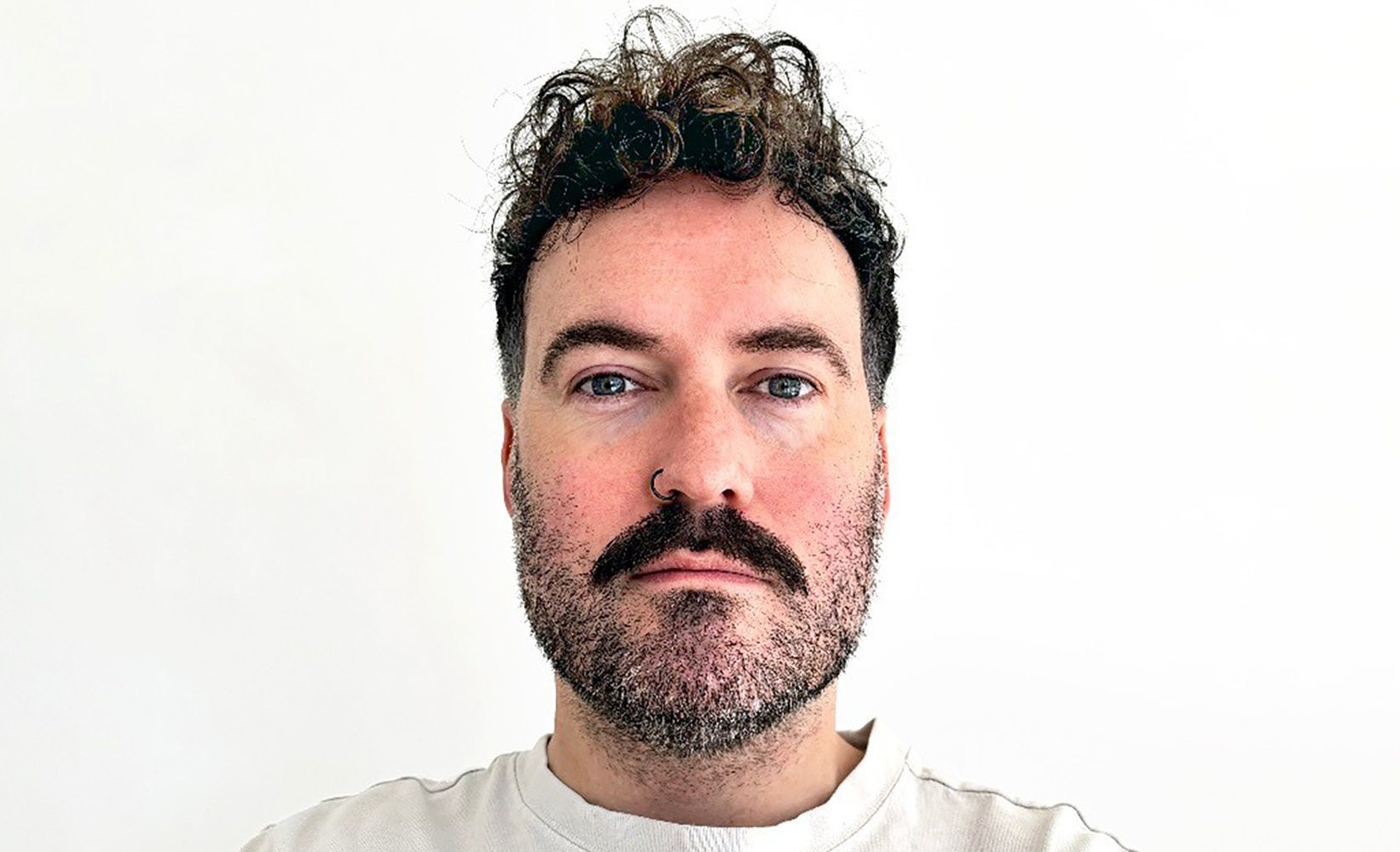 Man with dark curly hair, full beard, and moustache wearing white t-shirt against white background, looking directly at camera.