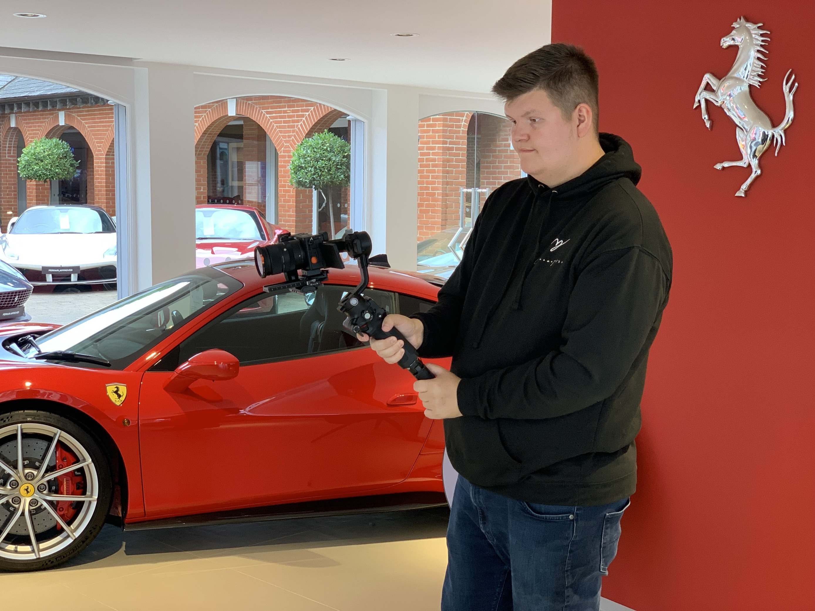 BA (Hons) Visual Effects graduate J McPherson holding a camera and tripod with a Ferrari logo and vehicle in the background.